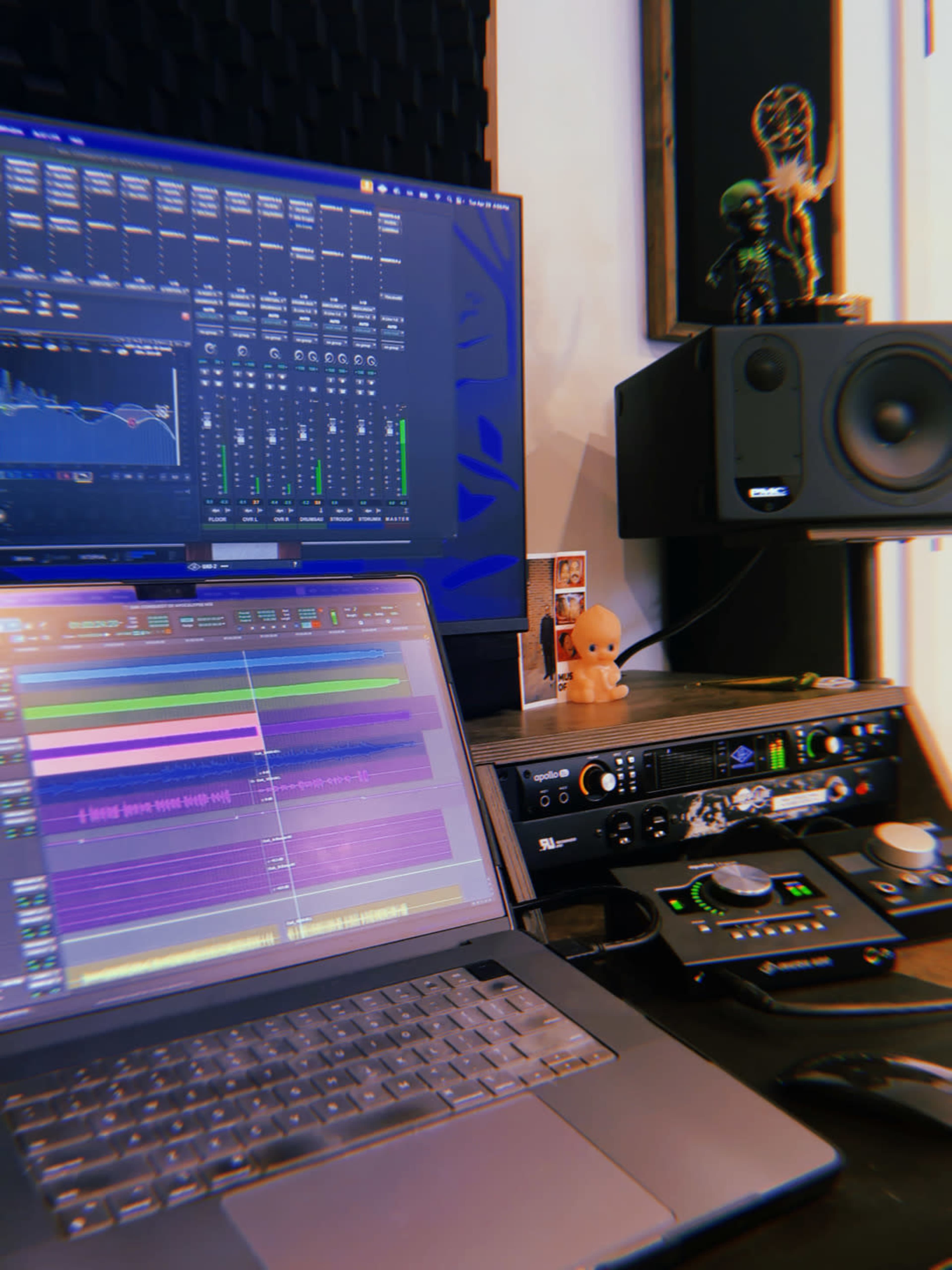 The image shows a music production setup with a laptop displaying digital audio tracks, a monitor with mixing software, and audio equipment on a desk.