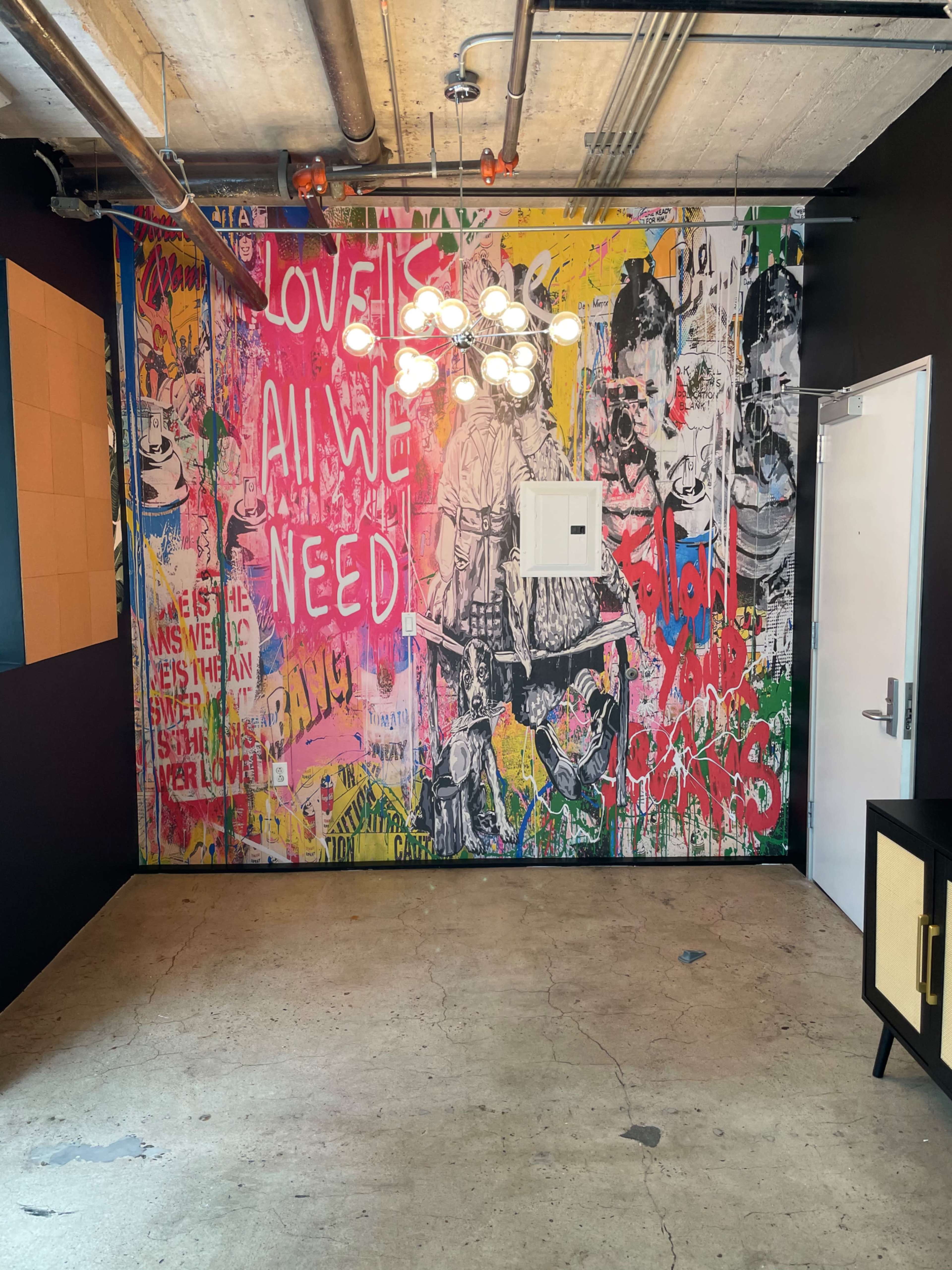 The image shows a room featuring a colorful, graffiti-covered wall with a light fixture and a door on the right.