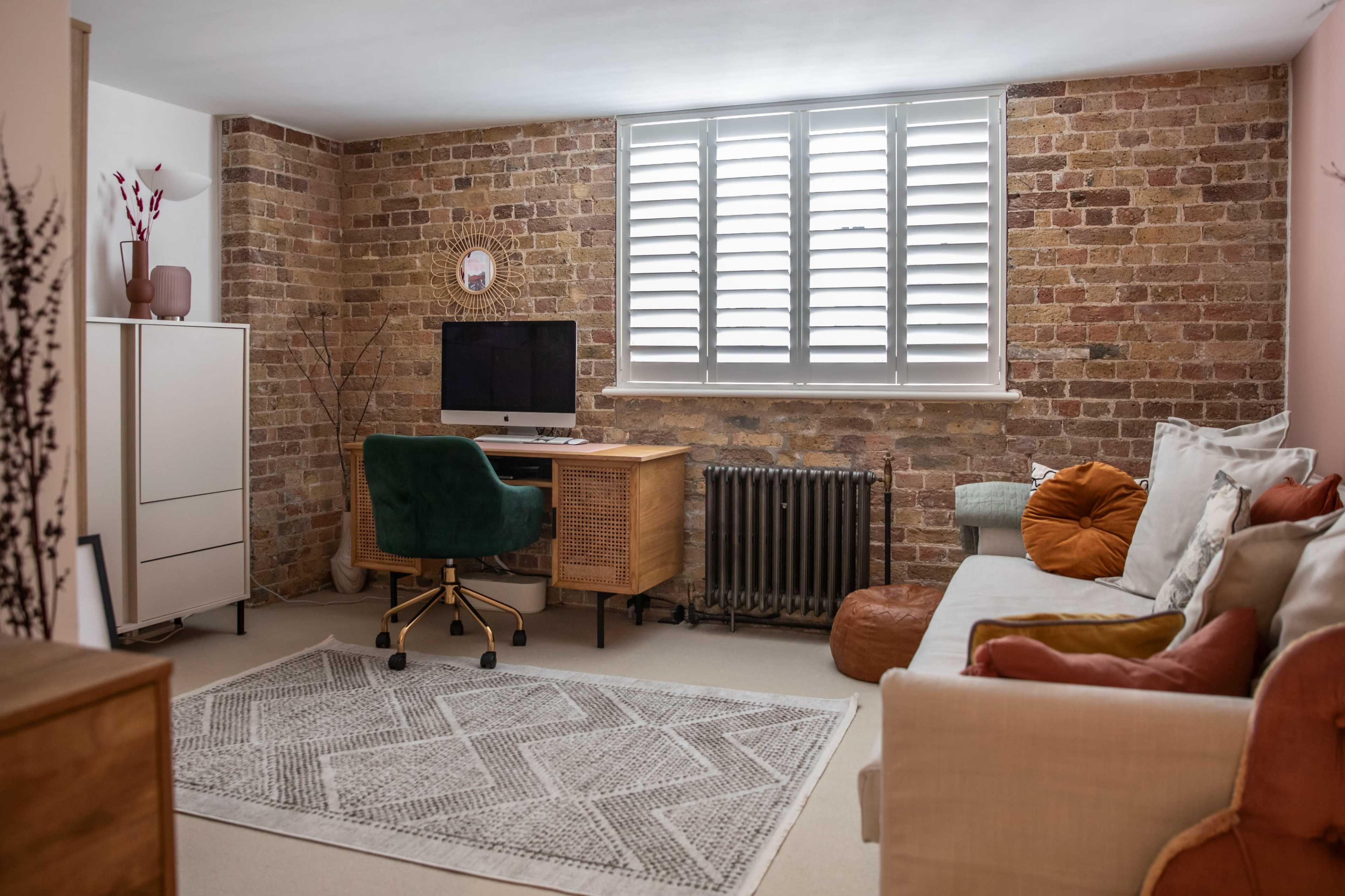 A modern workspace is set up against a brick wall, featuring a desk with a computer and a green chair, alongside a sofa and decorative pillows.