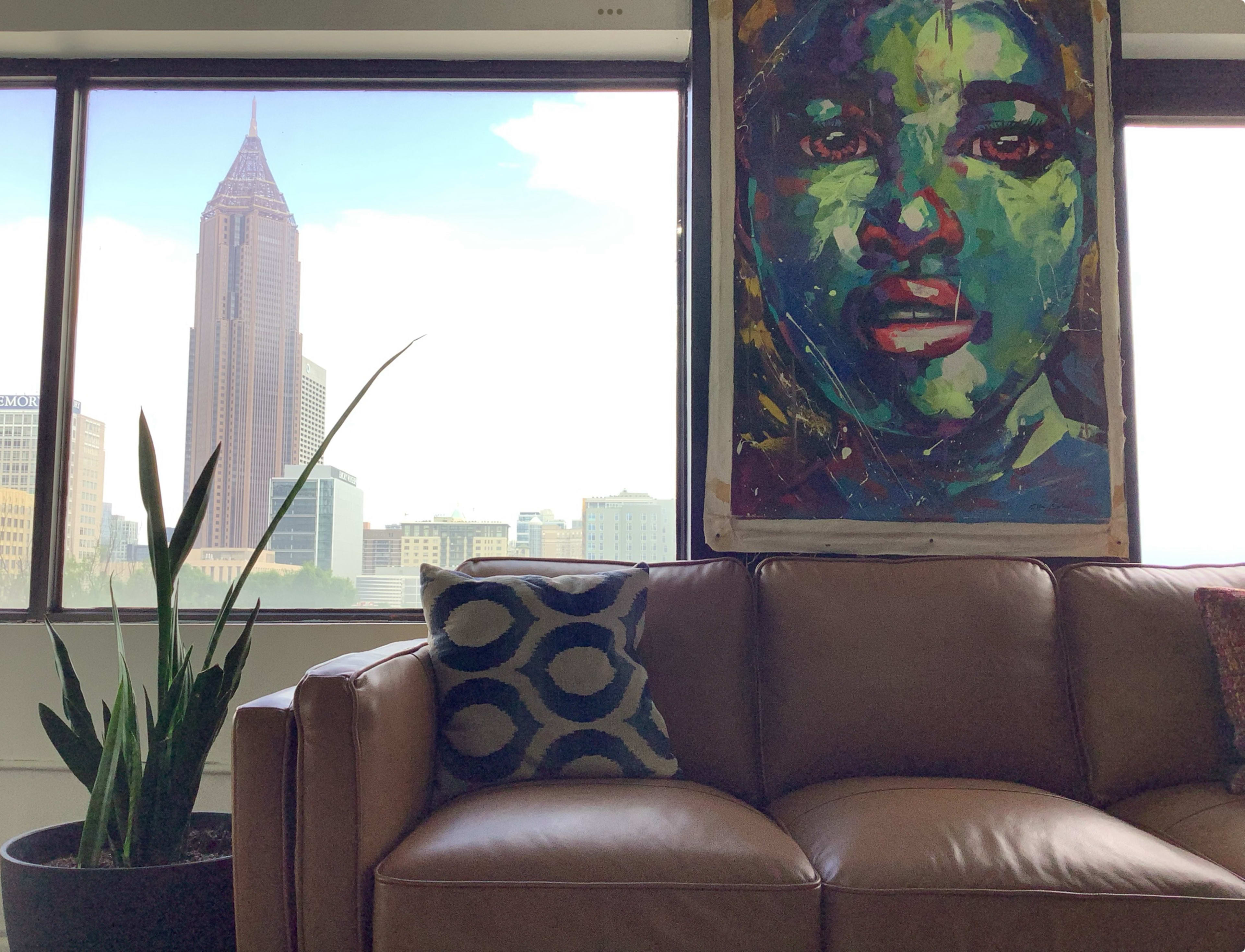 A large colorful portrait of a face hangs on the wall next to a brown leather couch, overlooking a cityscape that features a tall building with a pointed top.