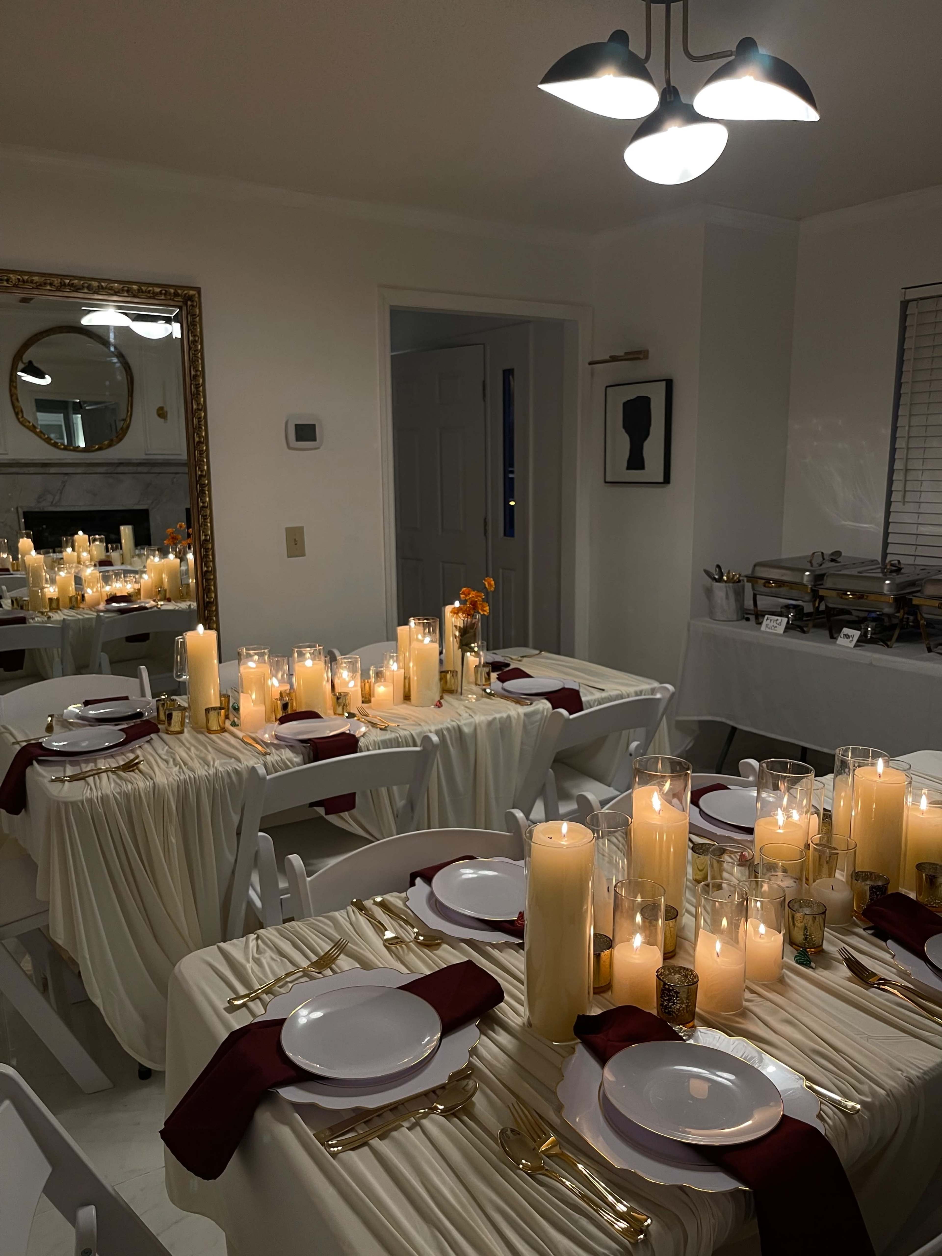 The image shows a dining room set for a formal dinner, featuring tables adorned with white tablecloths, maroon napkins, and numerous candles arranged in glass holders.