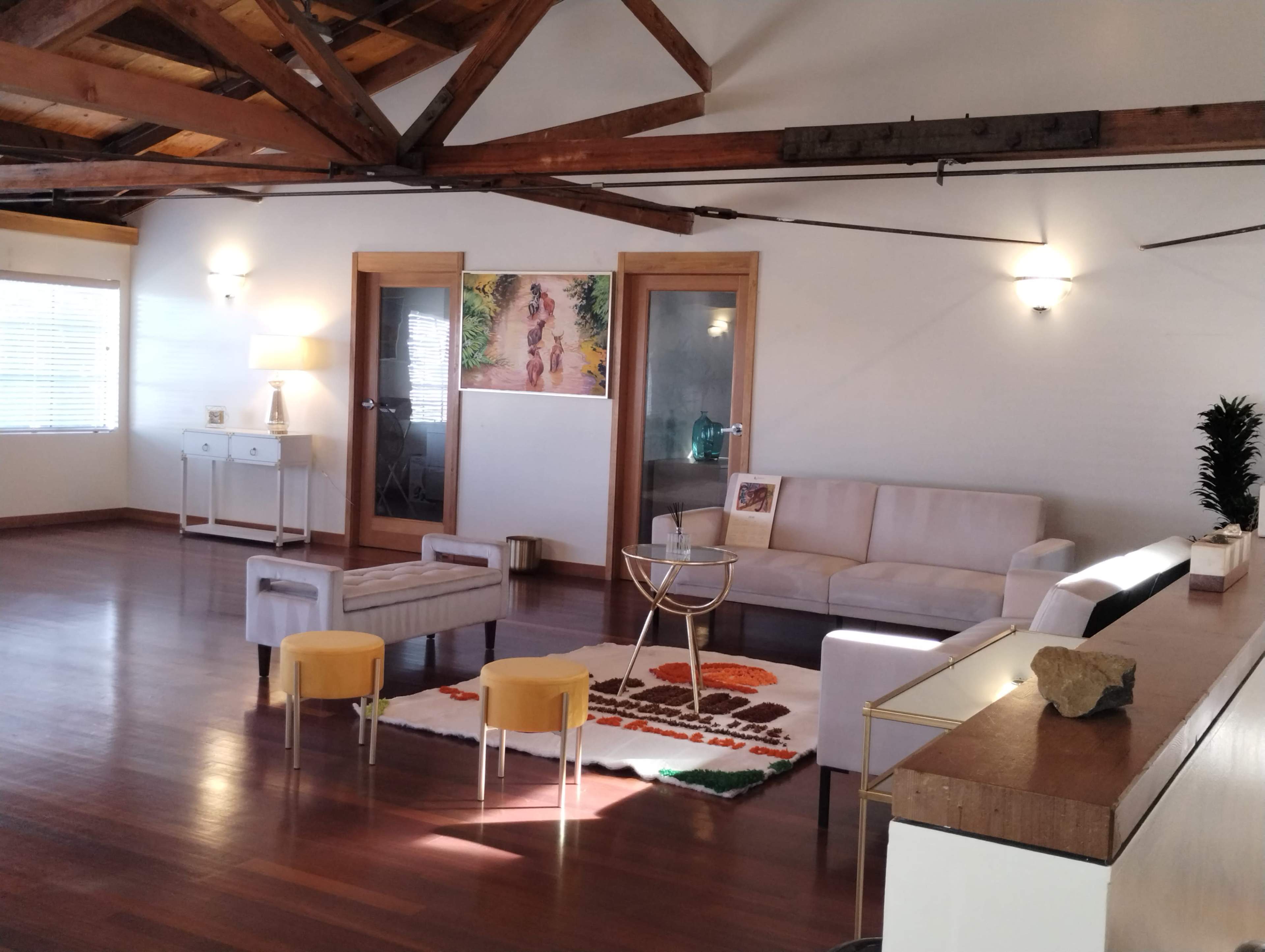 A spacious room with wooden beams, a light-colored sofa, a coffee table, and a decorative rug, featuring a side table and large windows.