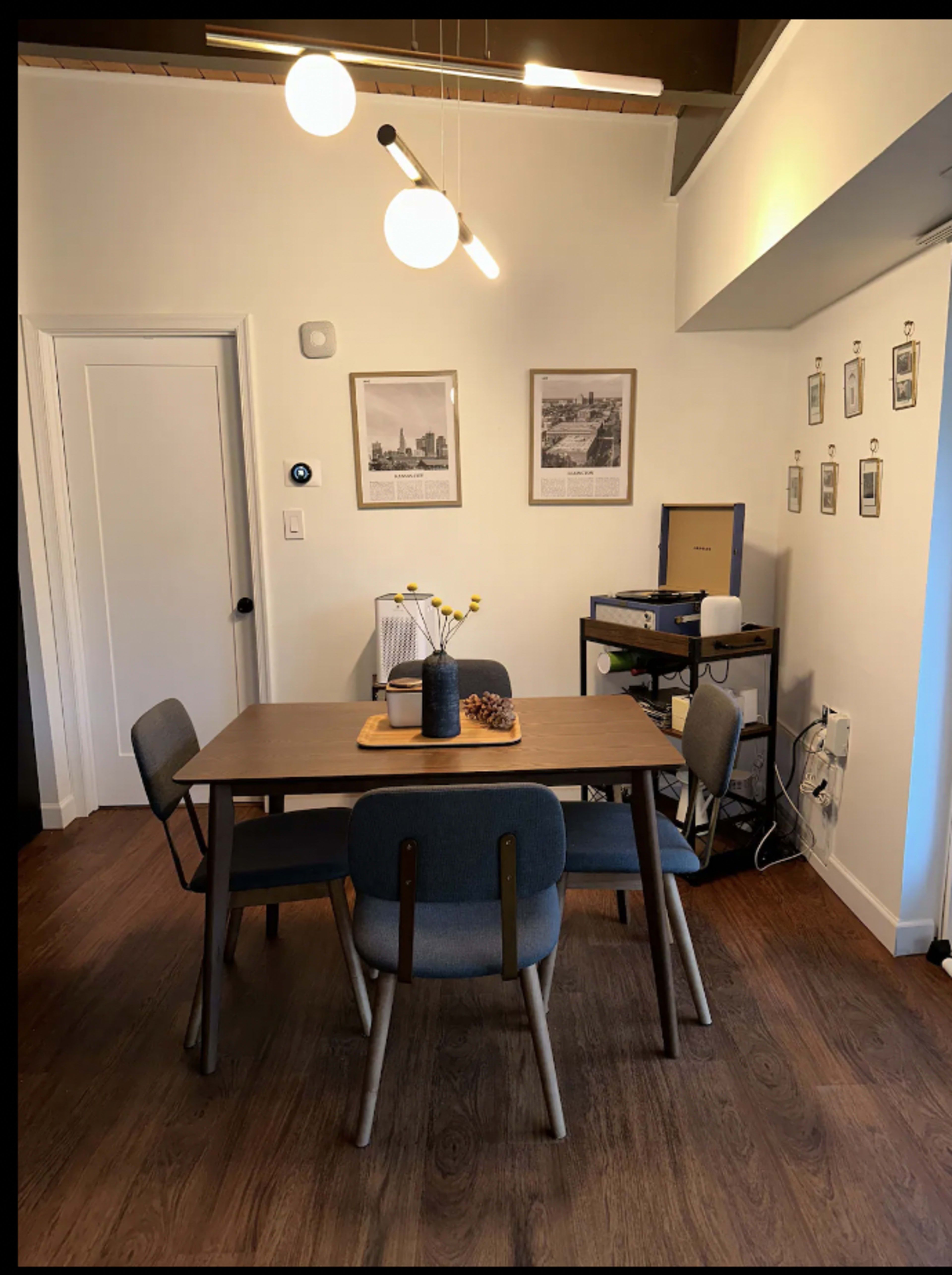 A dining area with a wooden table surrounded by four chairs, illuminated by a modern light fixture, and decorated with a vase of flowers and framed pictures on the walls.