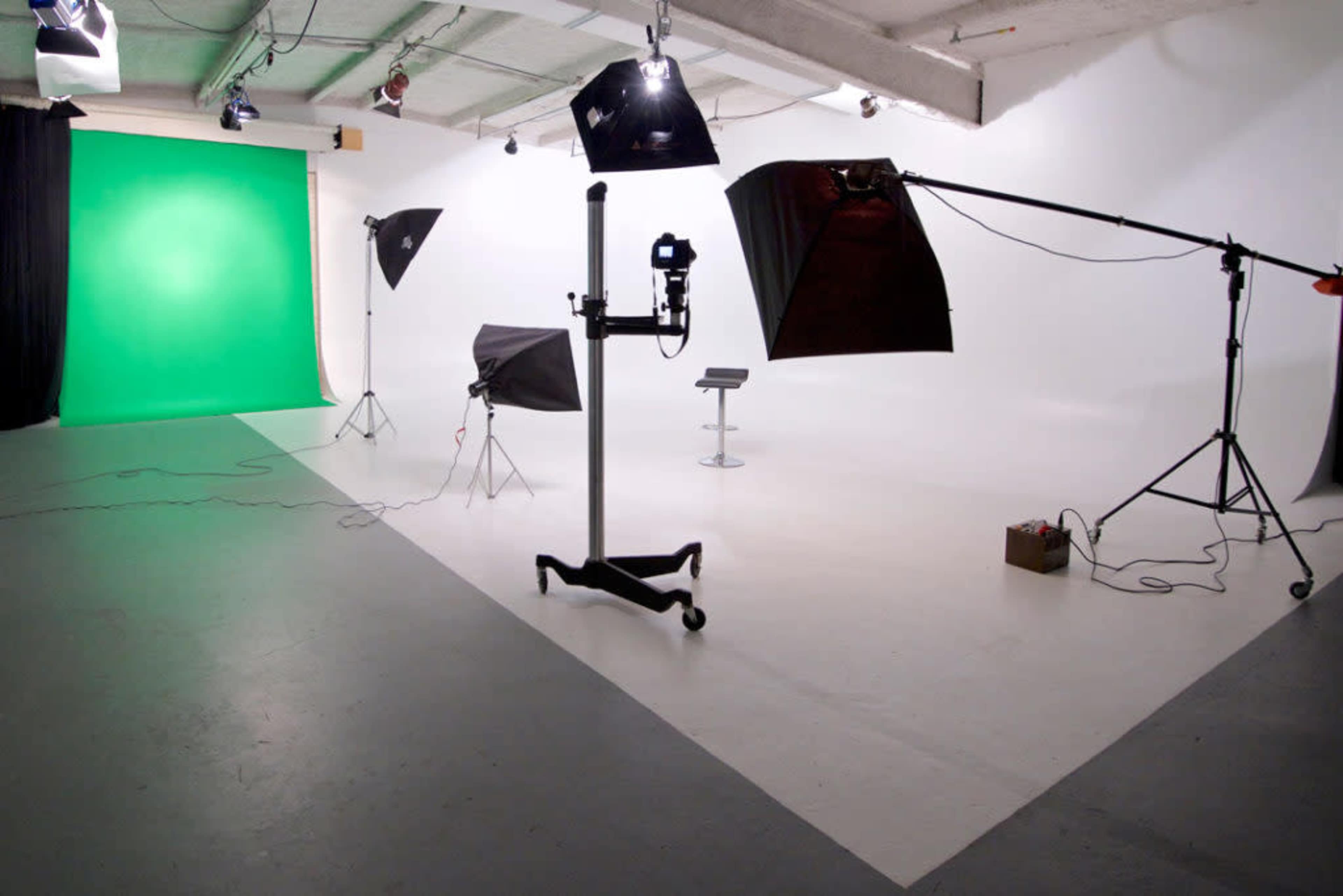 A photography studio with a green backdrop, lighting equipment, and a small stool in a corner.