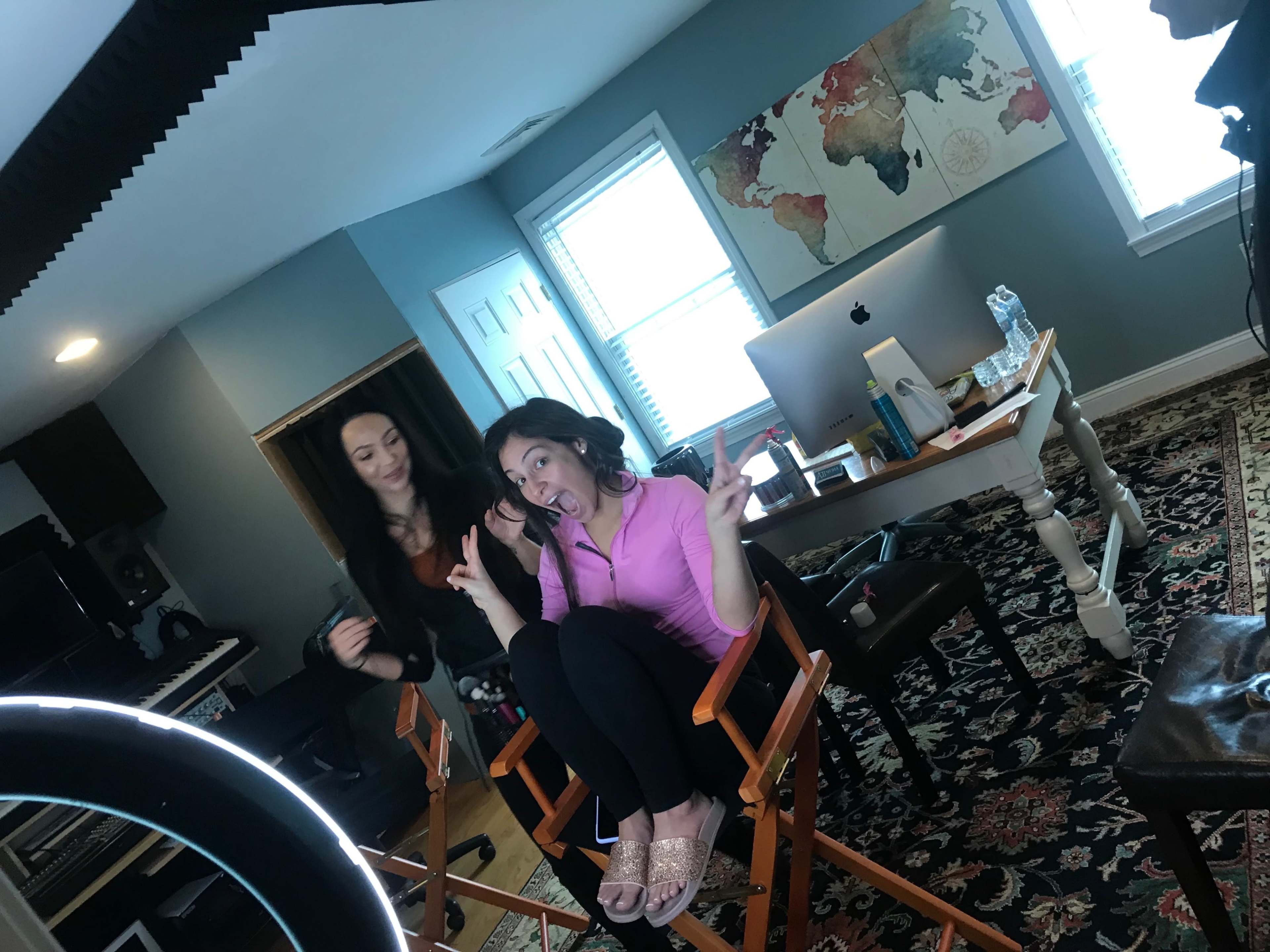 A woman in a pink shirt sits in a director's chair making a playful gesture while another woman stands behind her in an office setting.