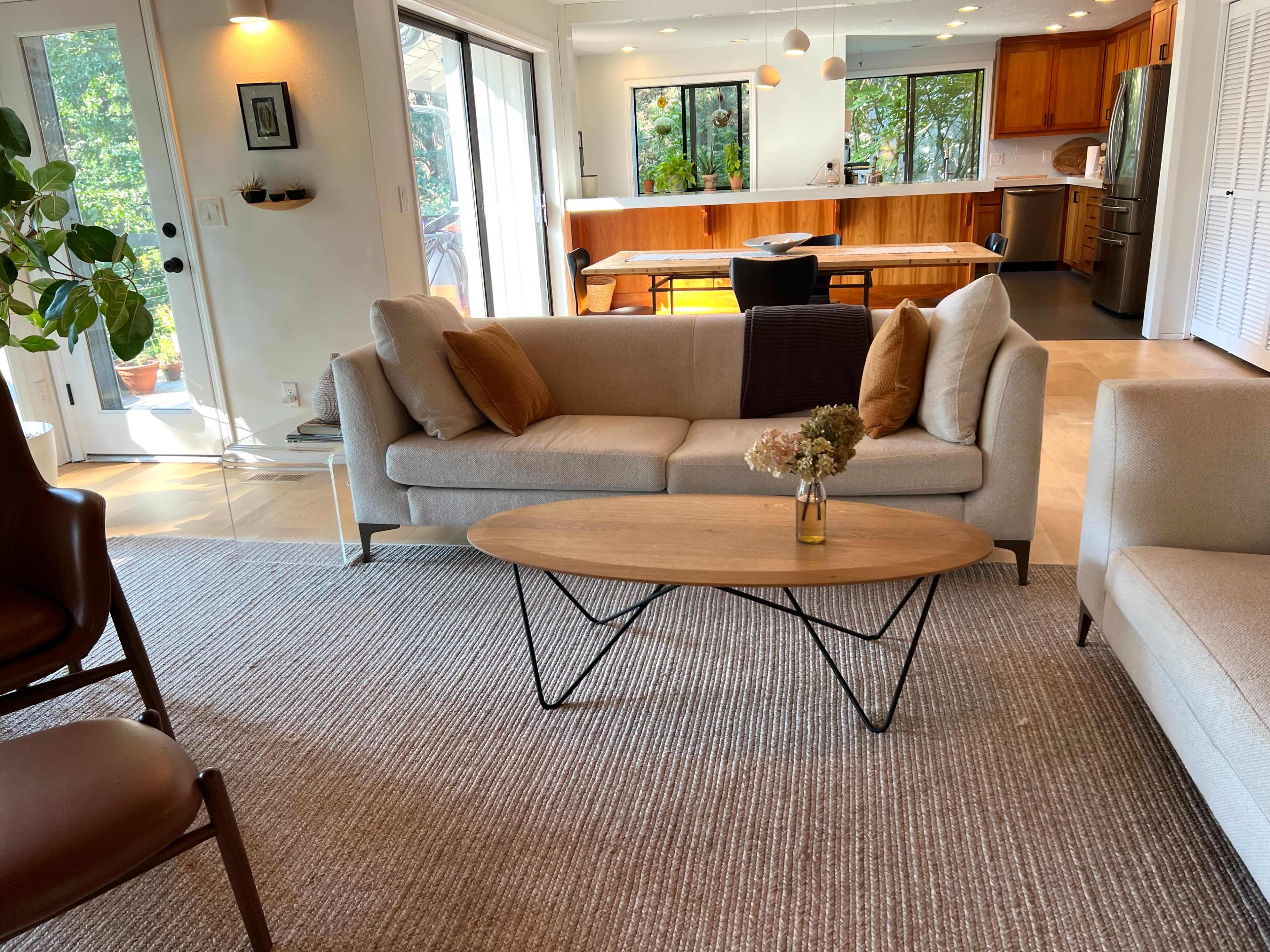 A living room features a light-colored couch and a wooden coffee table on a textured rug, with a view of a dining area and kitchen in the background.