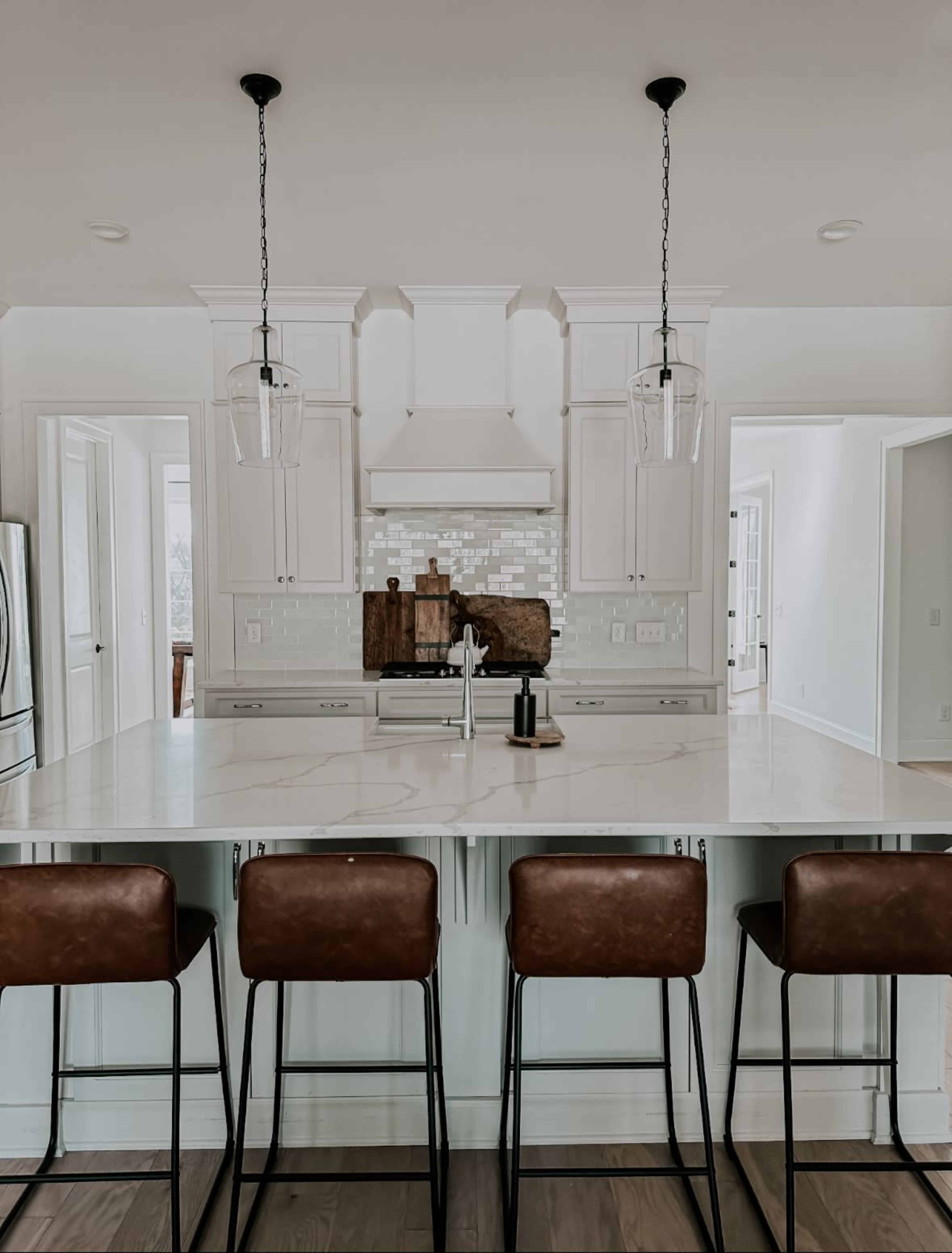 A modern kitchen features a large island with four leather bar stools, pendant lighting, and white cabinetry.