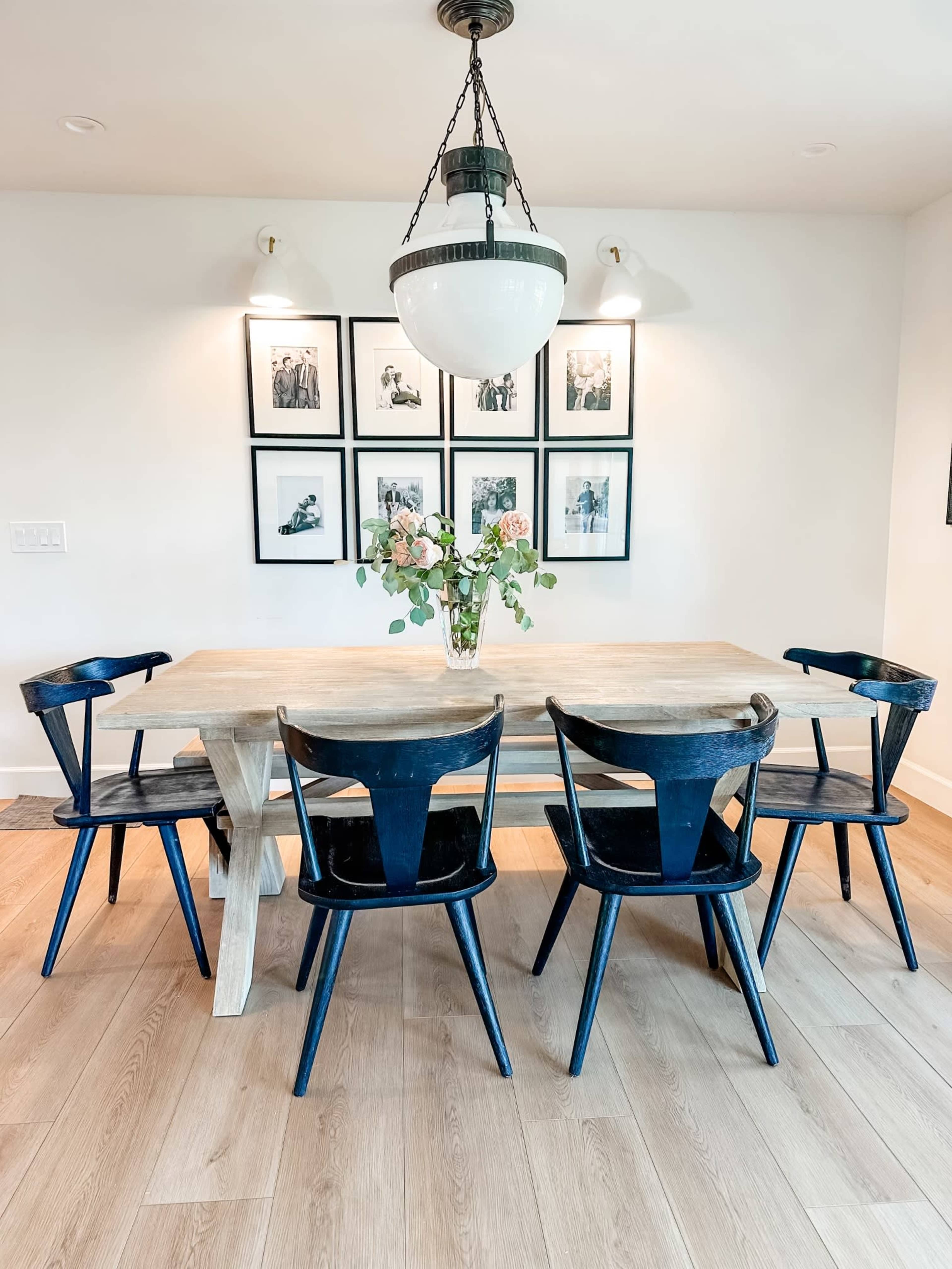A wooden dining table with a vase of flowers is surrounded by four black chairs, with framed photographs on the wall behind.