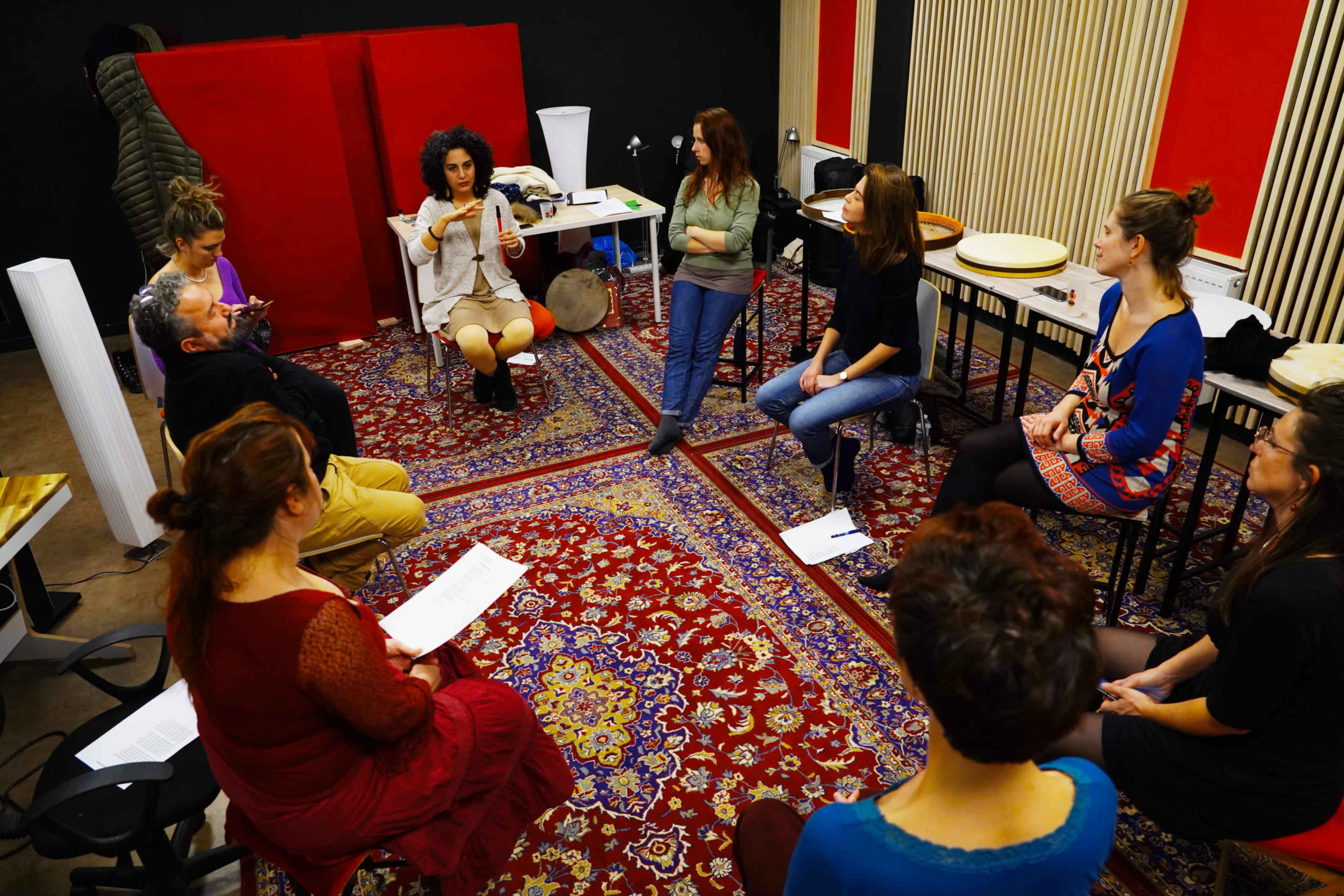 A group of people is seated in a circle on a patterned rug in a studio, engaging in a discussion led by a woman in the center.