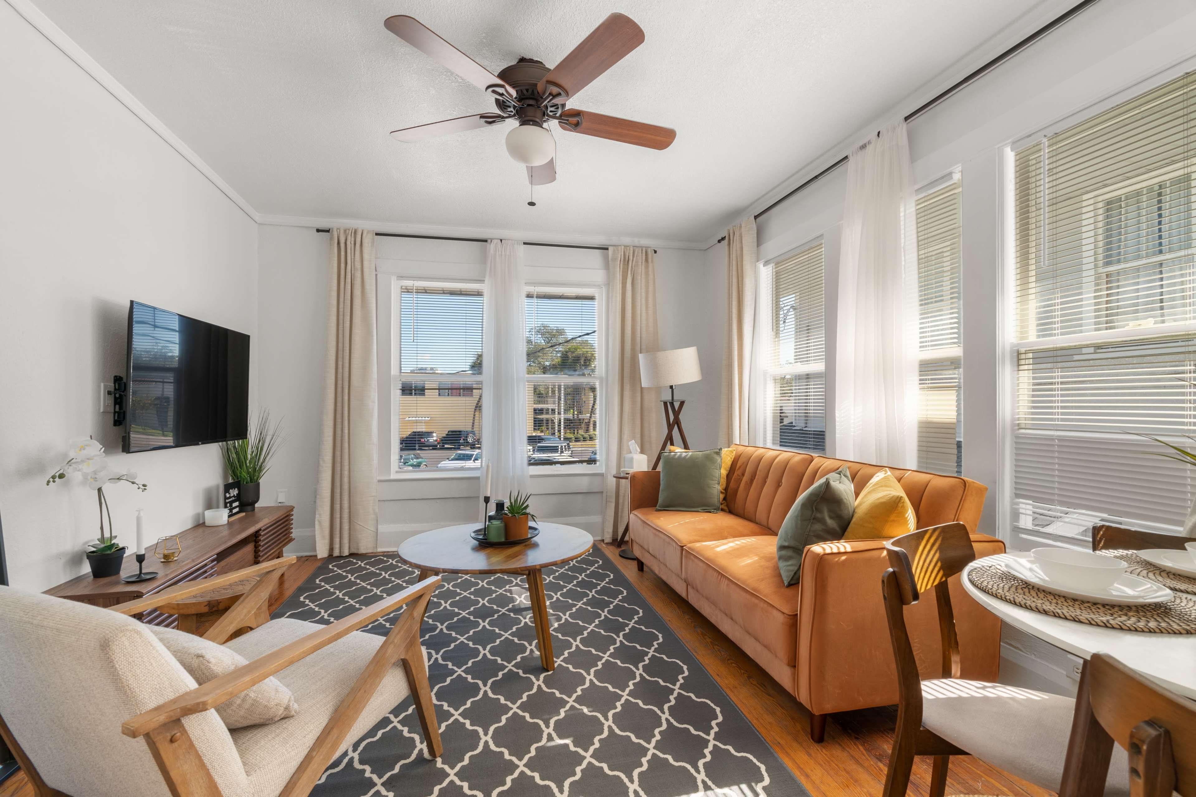 The living room features a tan sofa, light wood furniture, and large windows with sheer curtains, creating a bright and cozy space.