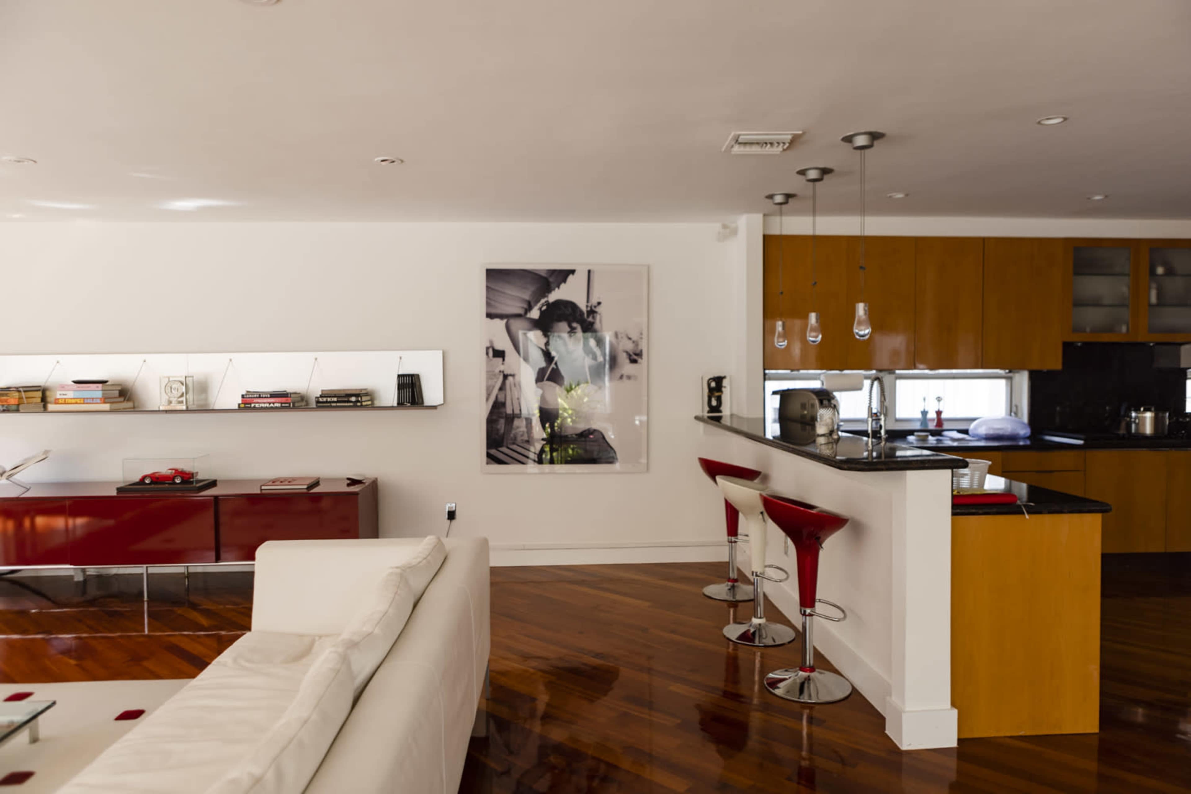 The image shows a modern kitchen and living area with a white sofa, red bar stools, and a large wall-mounted artwork.