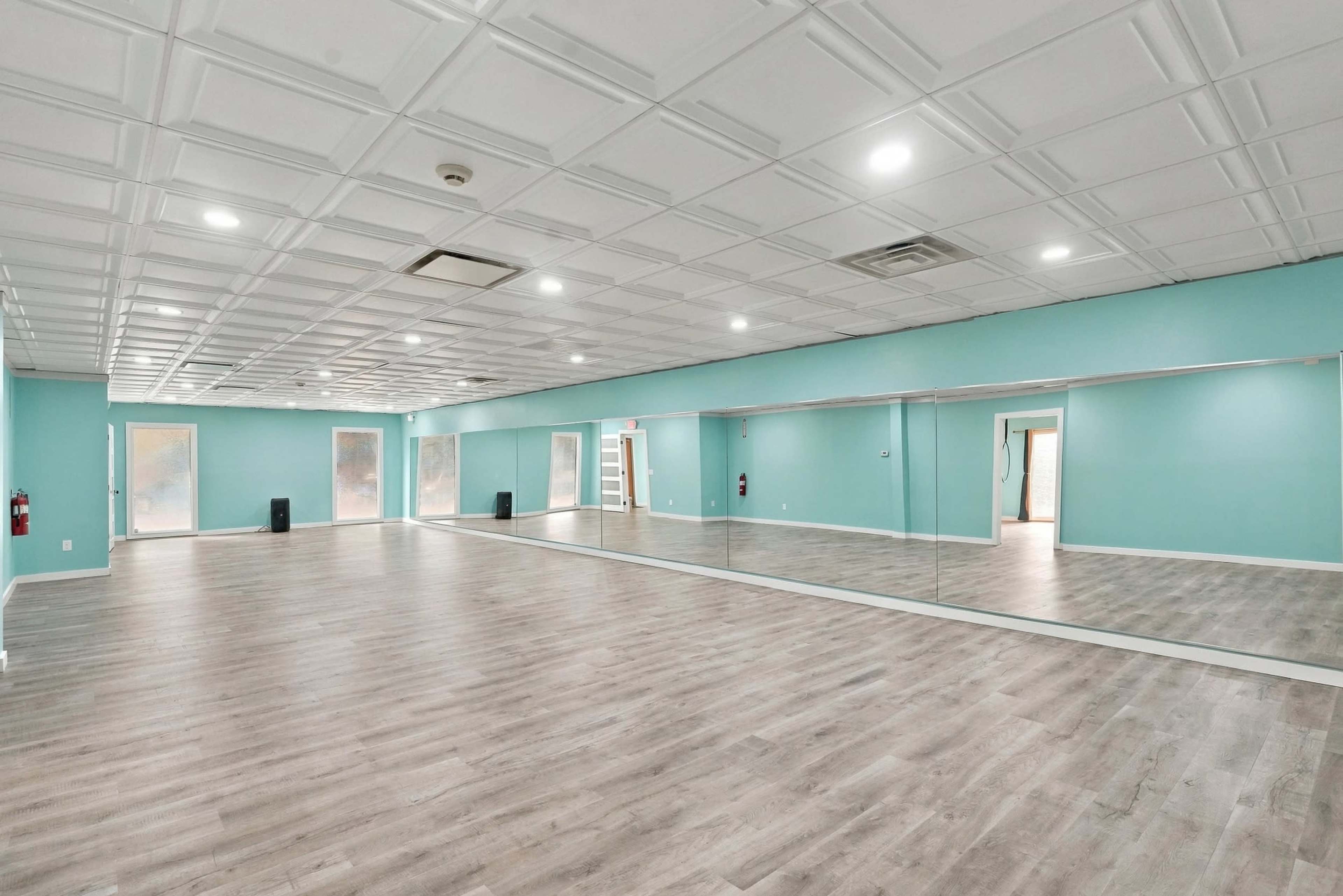 The image shows a spacious, well-lit room with light blue walls, large mirrors on one side, and a wooden floor.