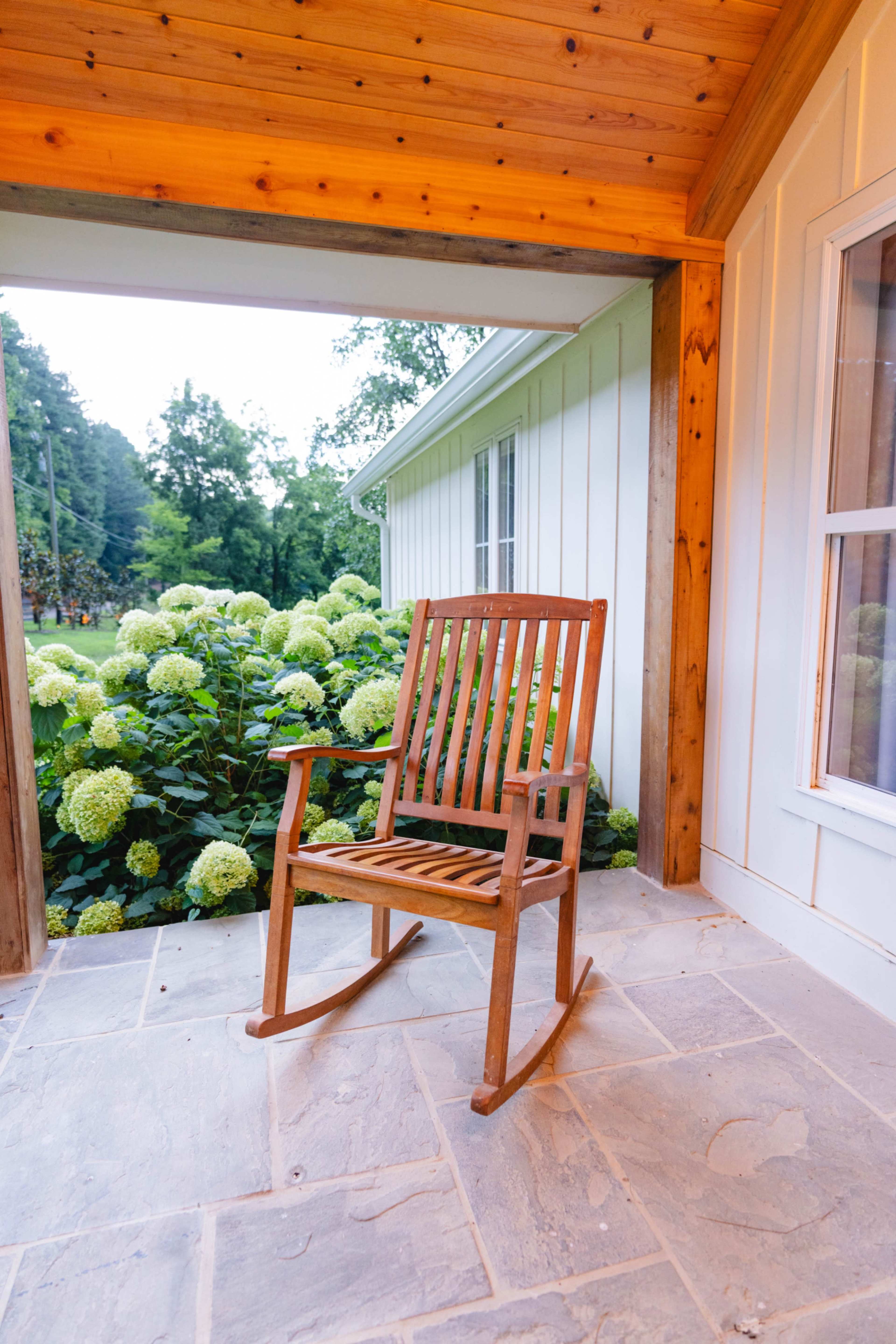 A wooden rocking chair is positioned on a stone porch in front of a blooming hydrangea bush.