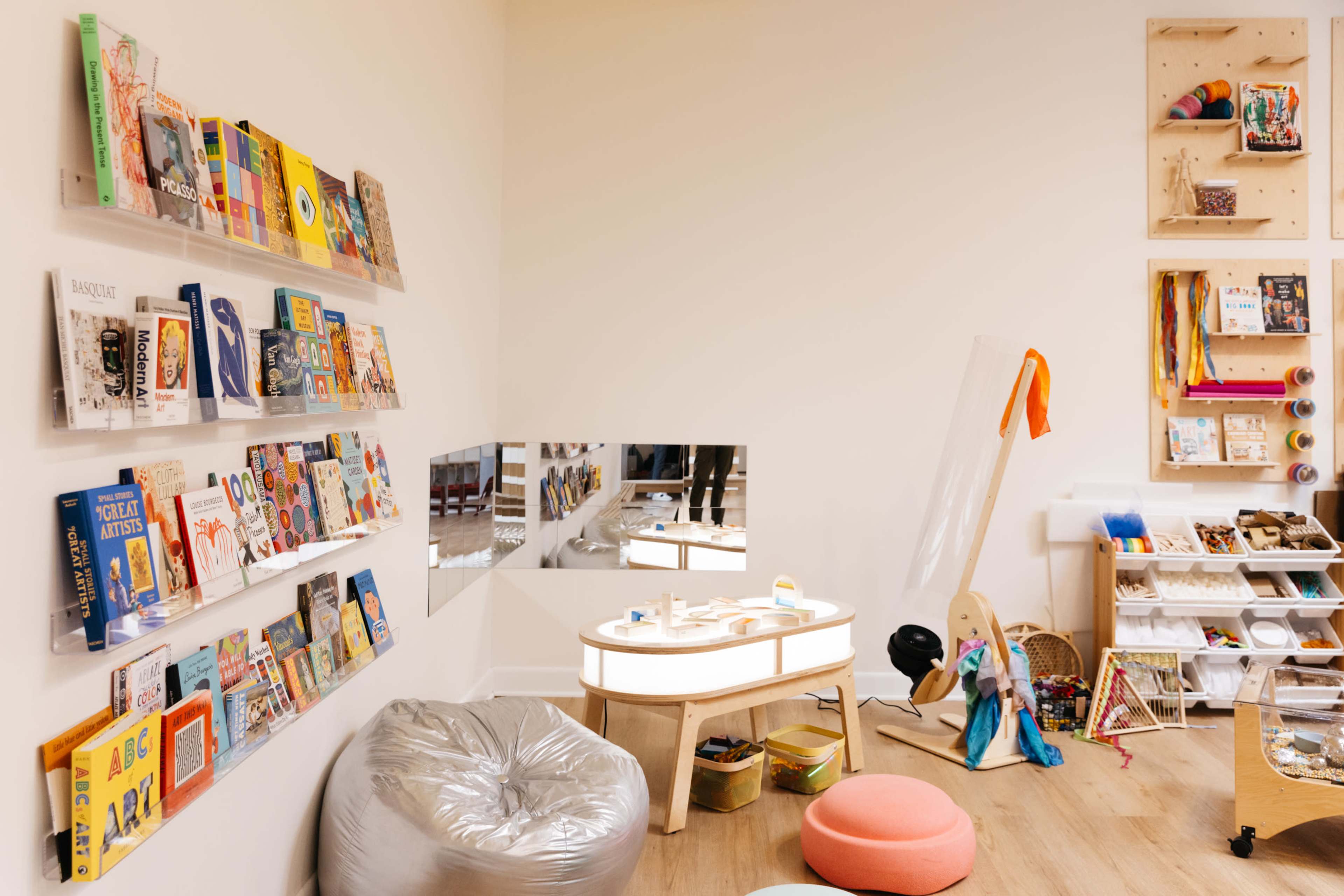 The image shows a bright, organized children's playroom featuring a wall of books, a mirrored play area, and various toys and materials scattered around.