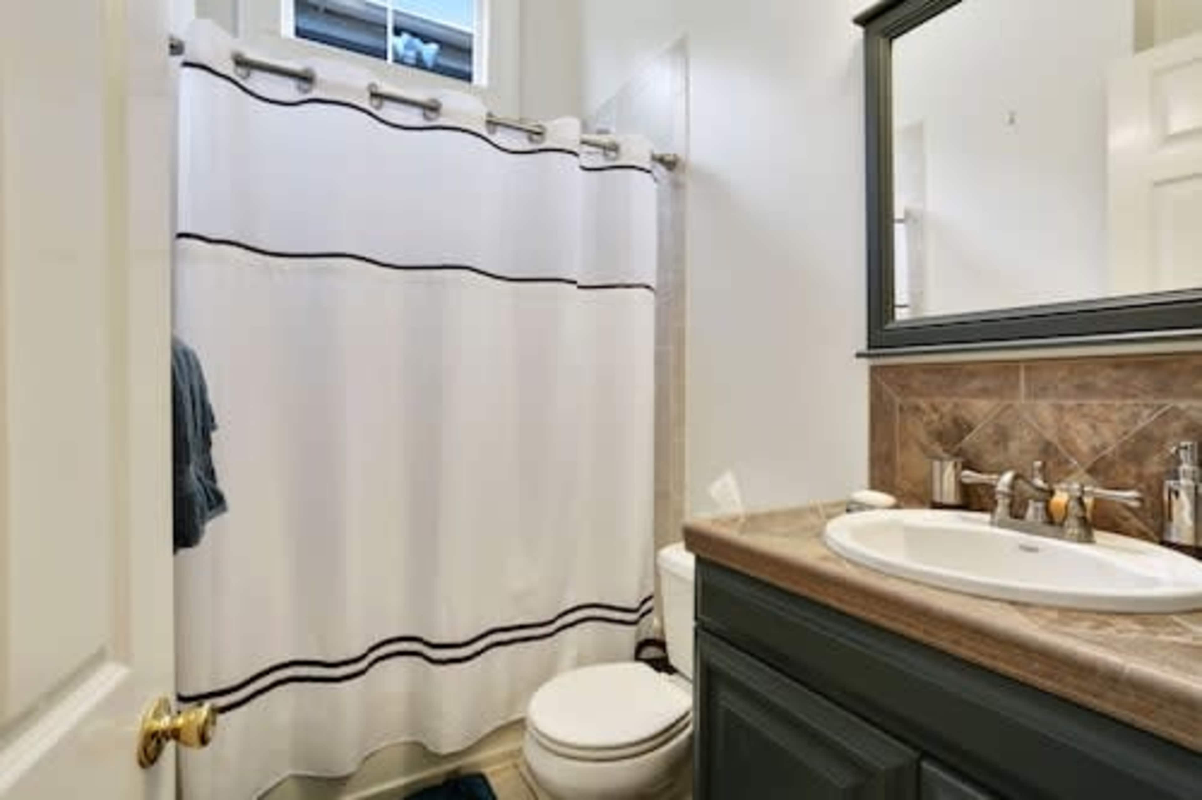 A bathroom featuring a shower with a white curtain, a sink with a mirror above it, and a toilet adjacent to the wall.