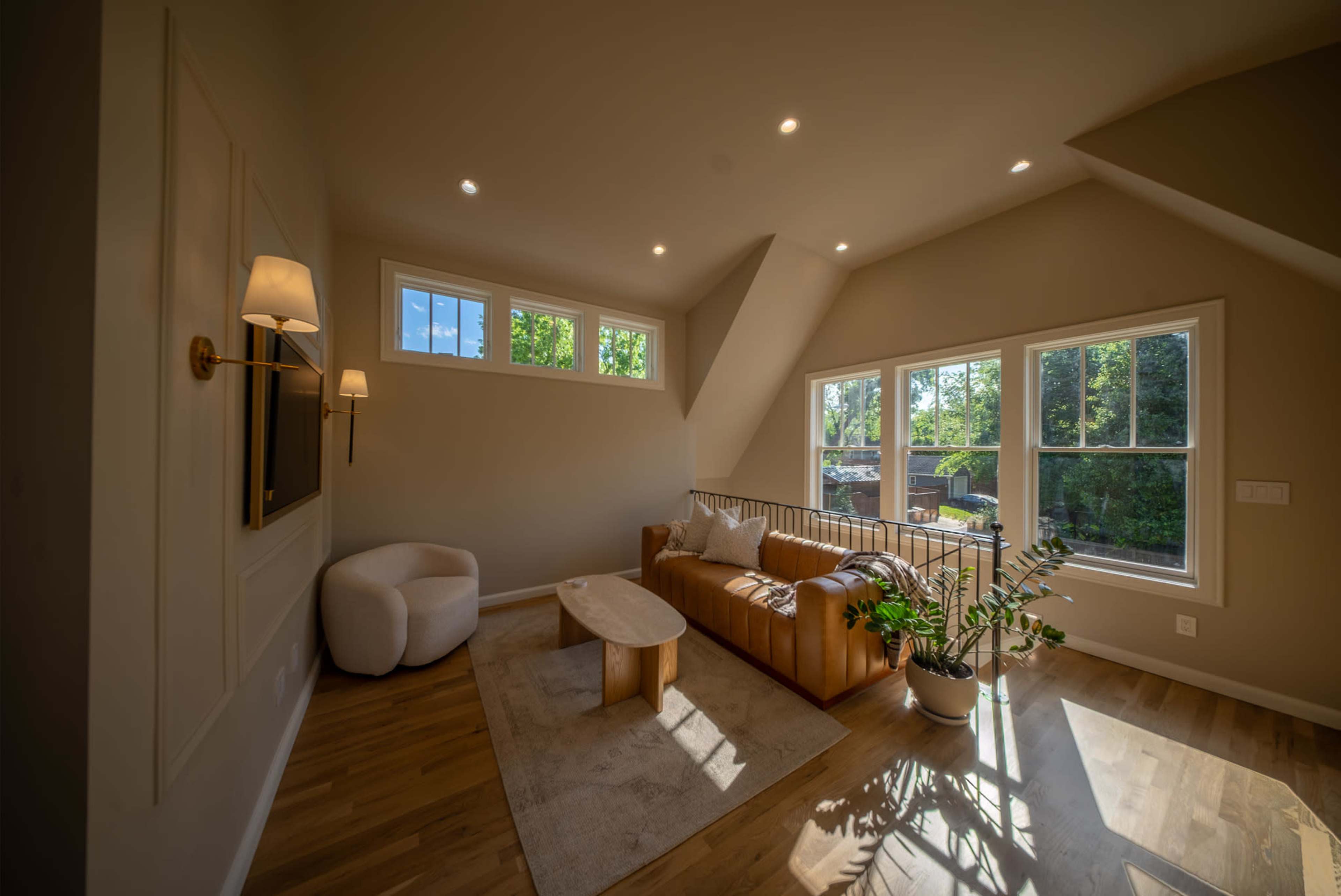 A cozy living room features a brown leather couch, a light wood coffee table, and large windows that allow natural light to fill the space.