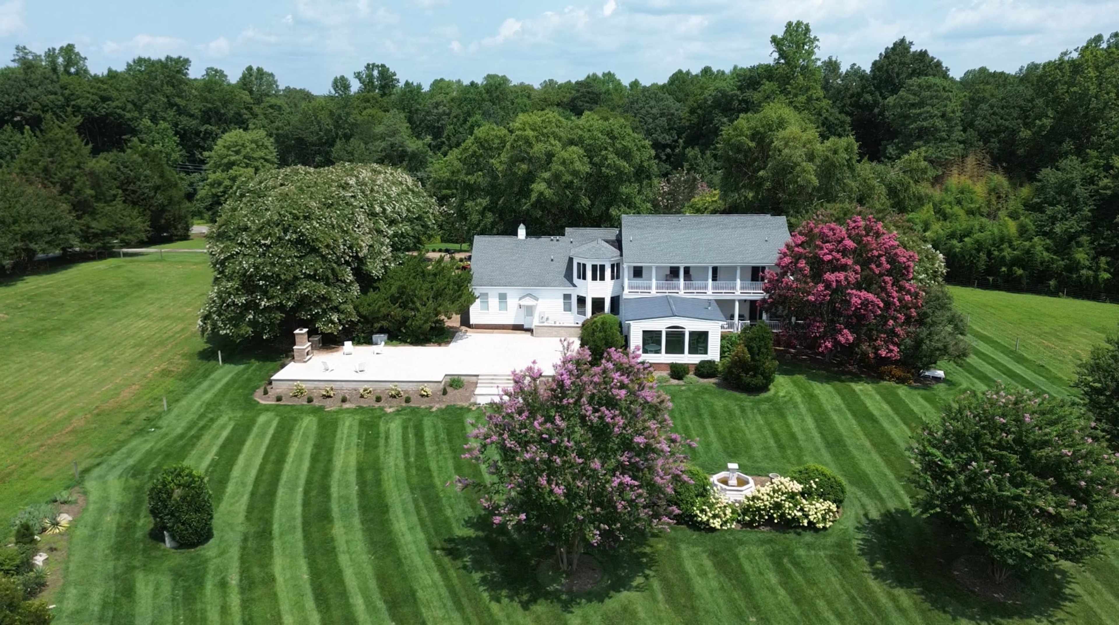 The image shows a large, two-story house surrounded by green lawns and flowering trees in a landscaped yard.