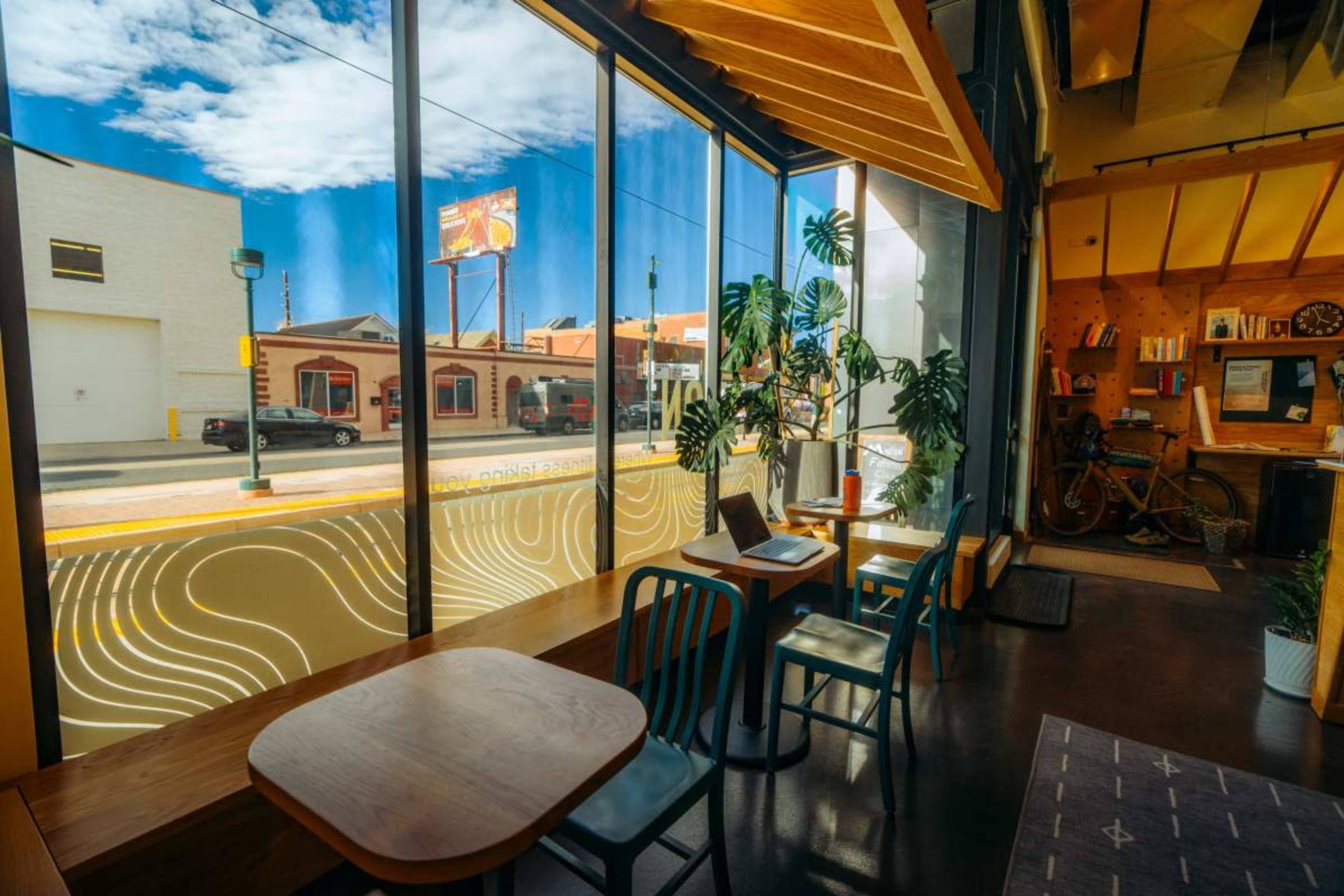 The image shows a brightly lit café with large windows, featuring two wooden tables and chairs, and a view of a street with buildings and a billboard outside.