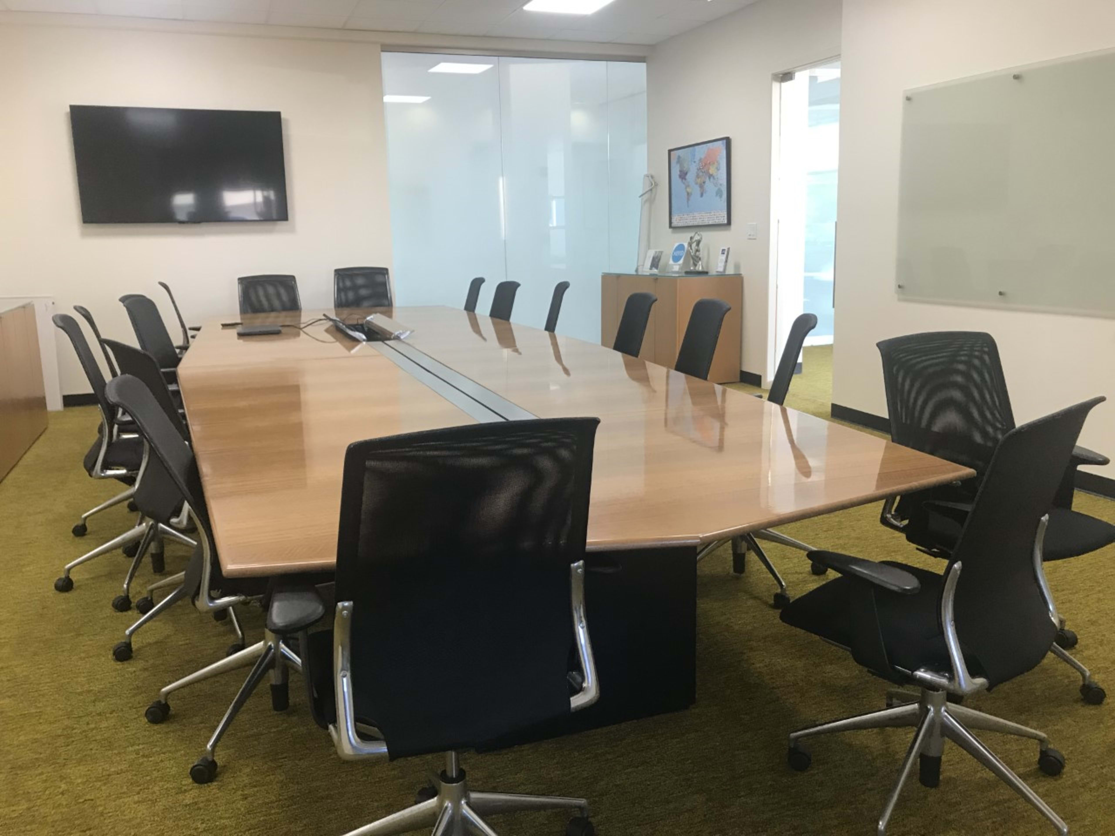 A large conference room features a rectangular wooden table surrounded by ergonomic black chairs, with a wall-mounted screen and a glass partition.