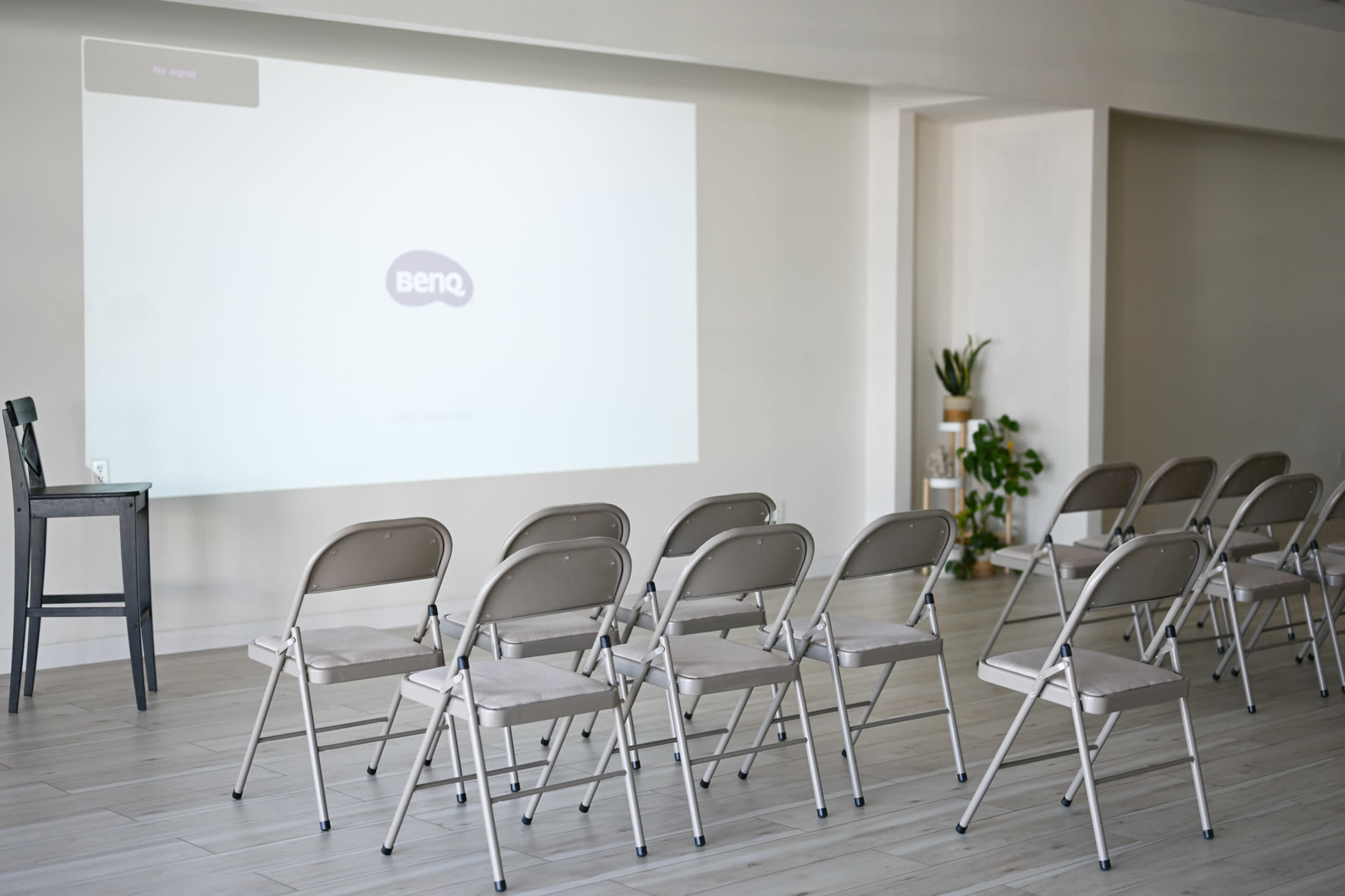 A plain room features several metal folding chairs arranged in rows facing a large blank projection screen.