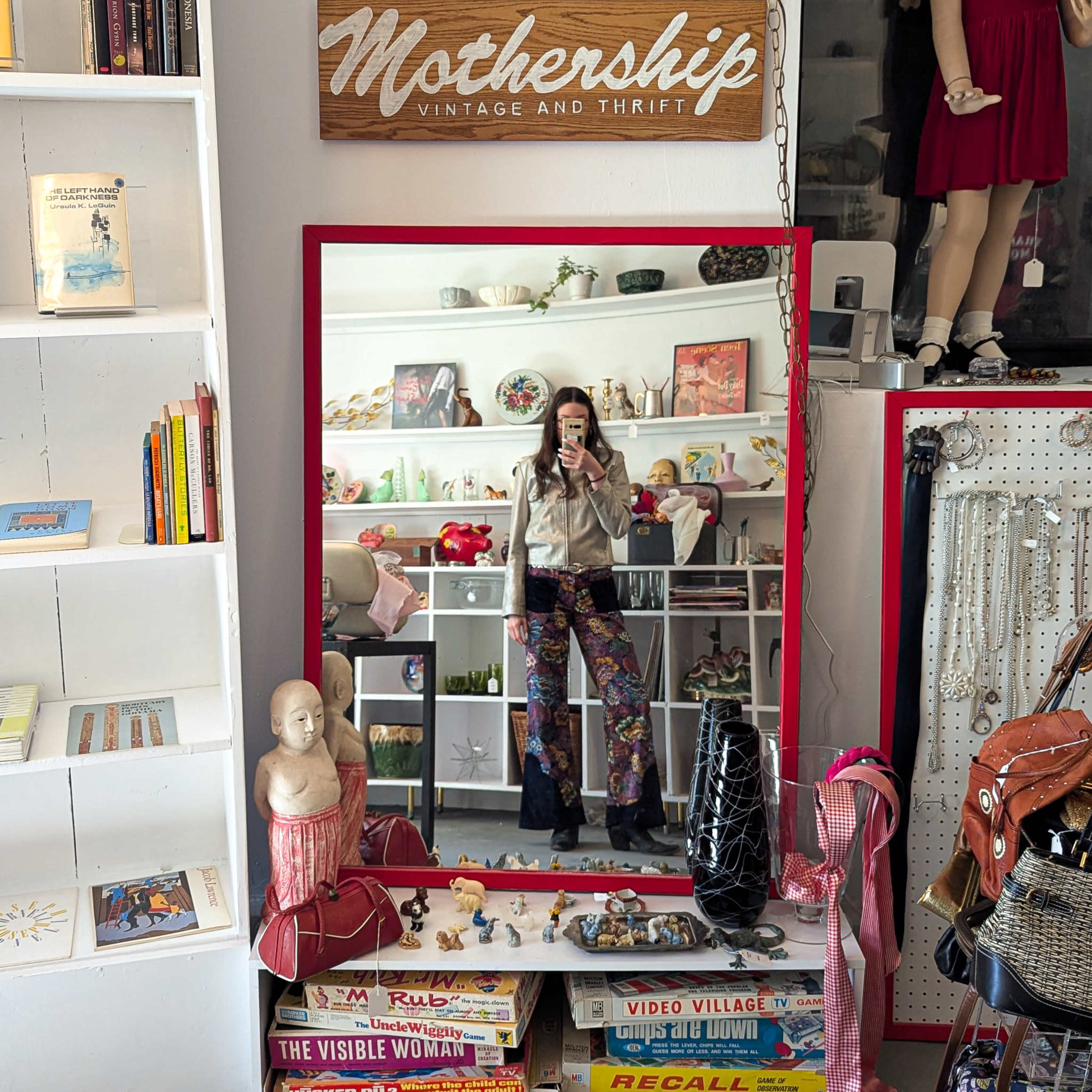 A person stands in front of a large mirror in a vintage shop, surrounded by various thrifted items and clothing.