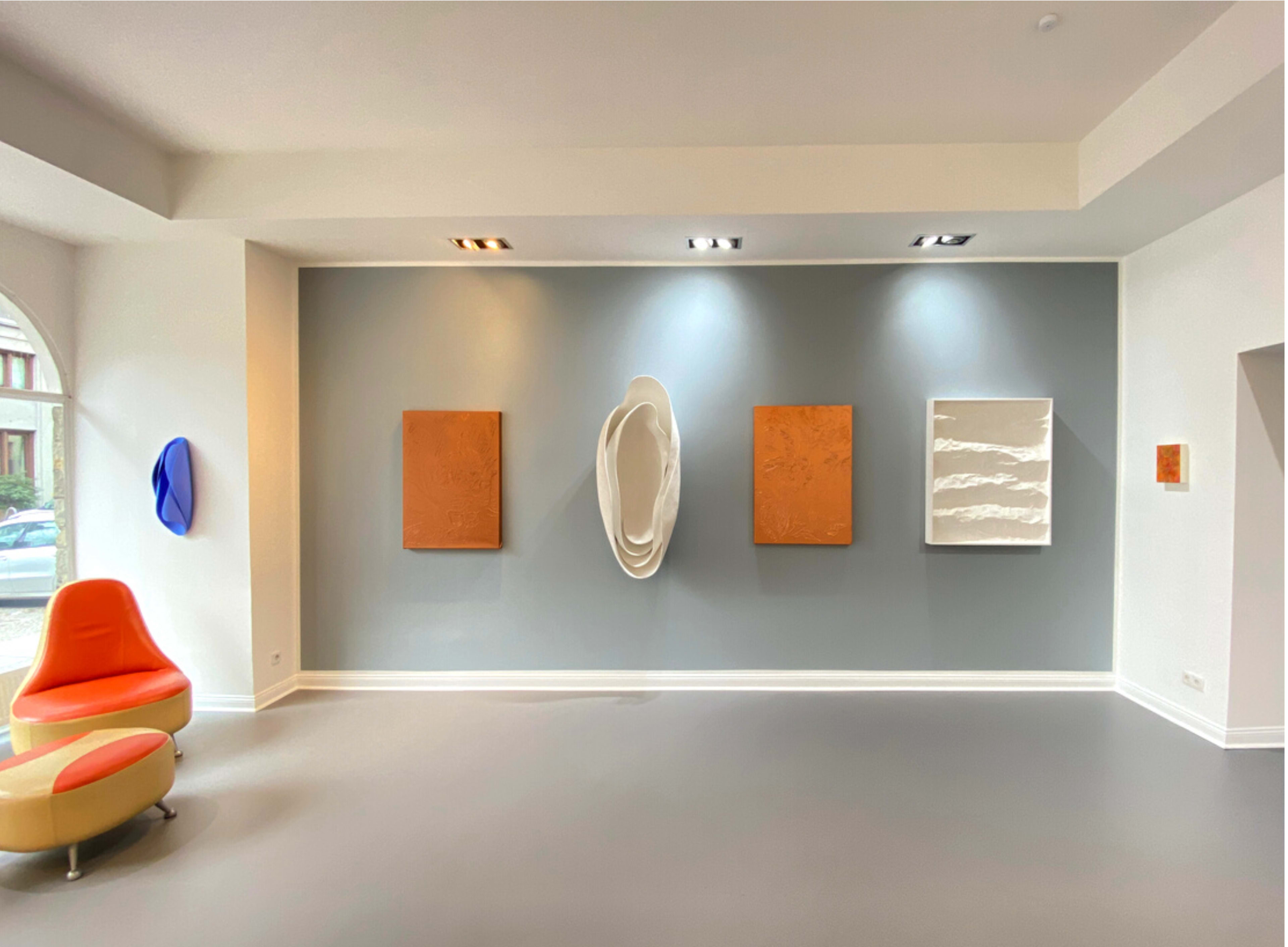 An art gallery featuring four abstract paintings on a gray wall, with a modern orange and yellow seating area in the foreground.