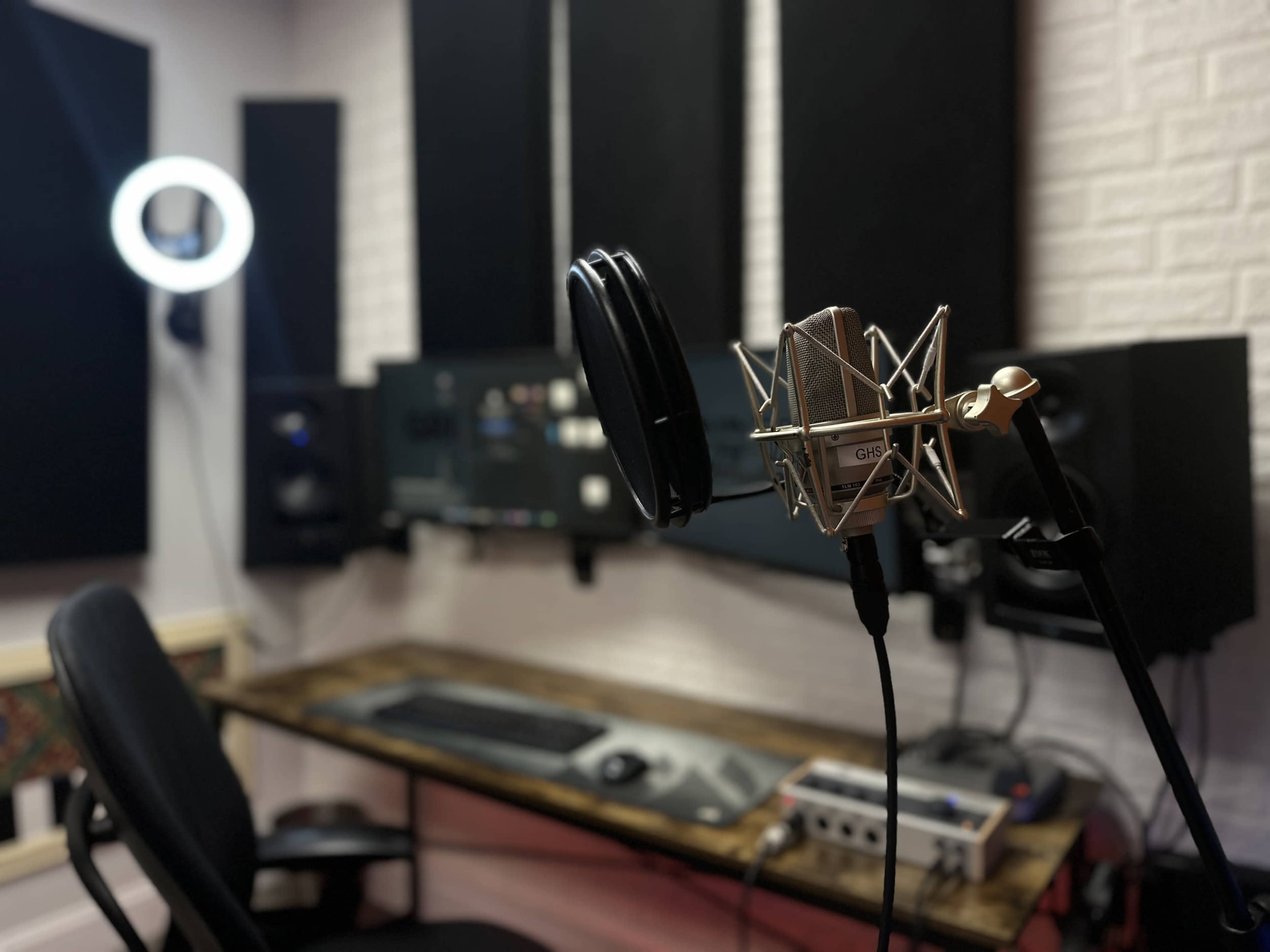 A microphone on a stand in a recording studio, with audio equipment and a desk visible in the background.
