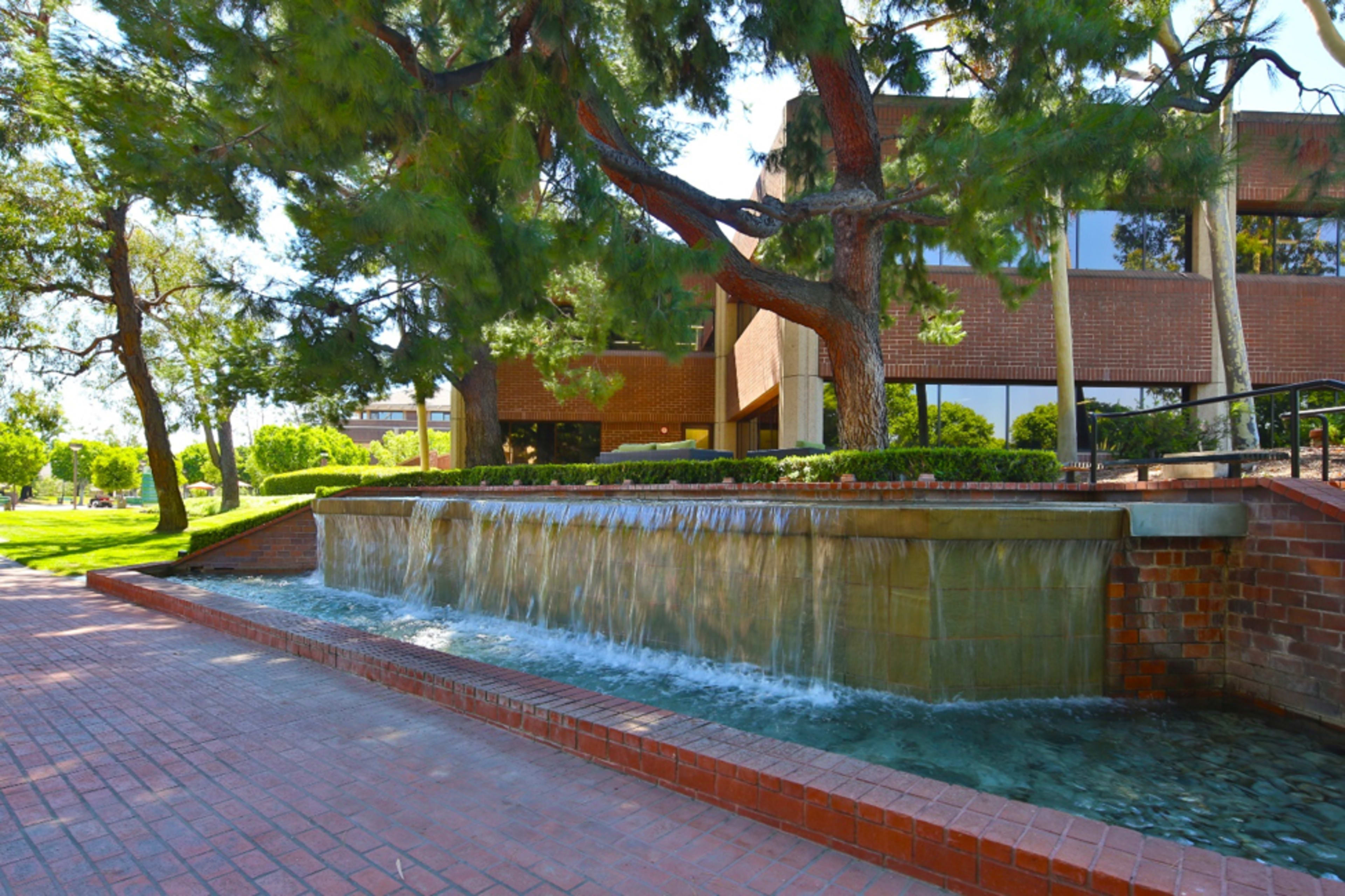 A low stone fountain with cascading water, surrounded by trees and a brick pathway adjacent to a modern building.