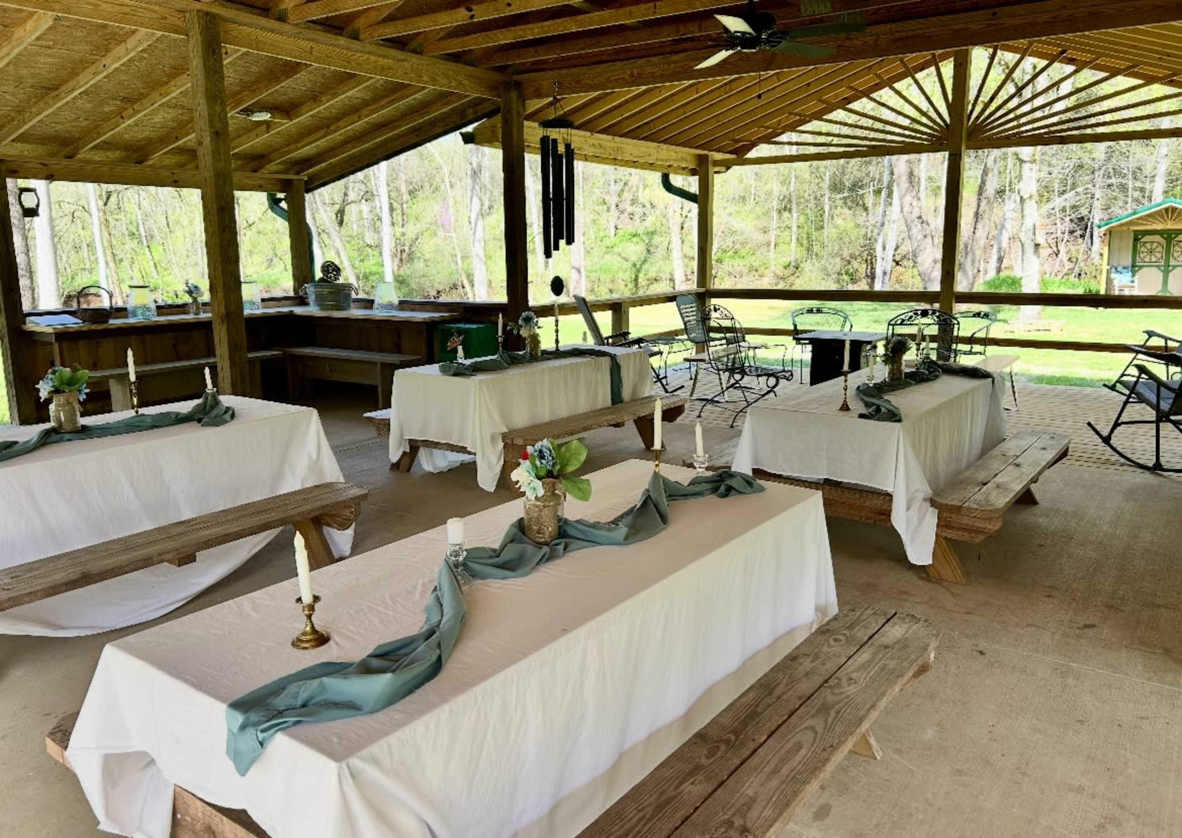 The image shows a covered outdoor area with several tables arranged with white tablecloths, each featuring a small decorative centerpiece and candles.