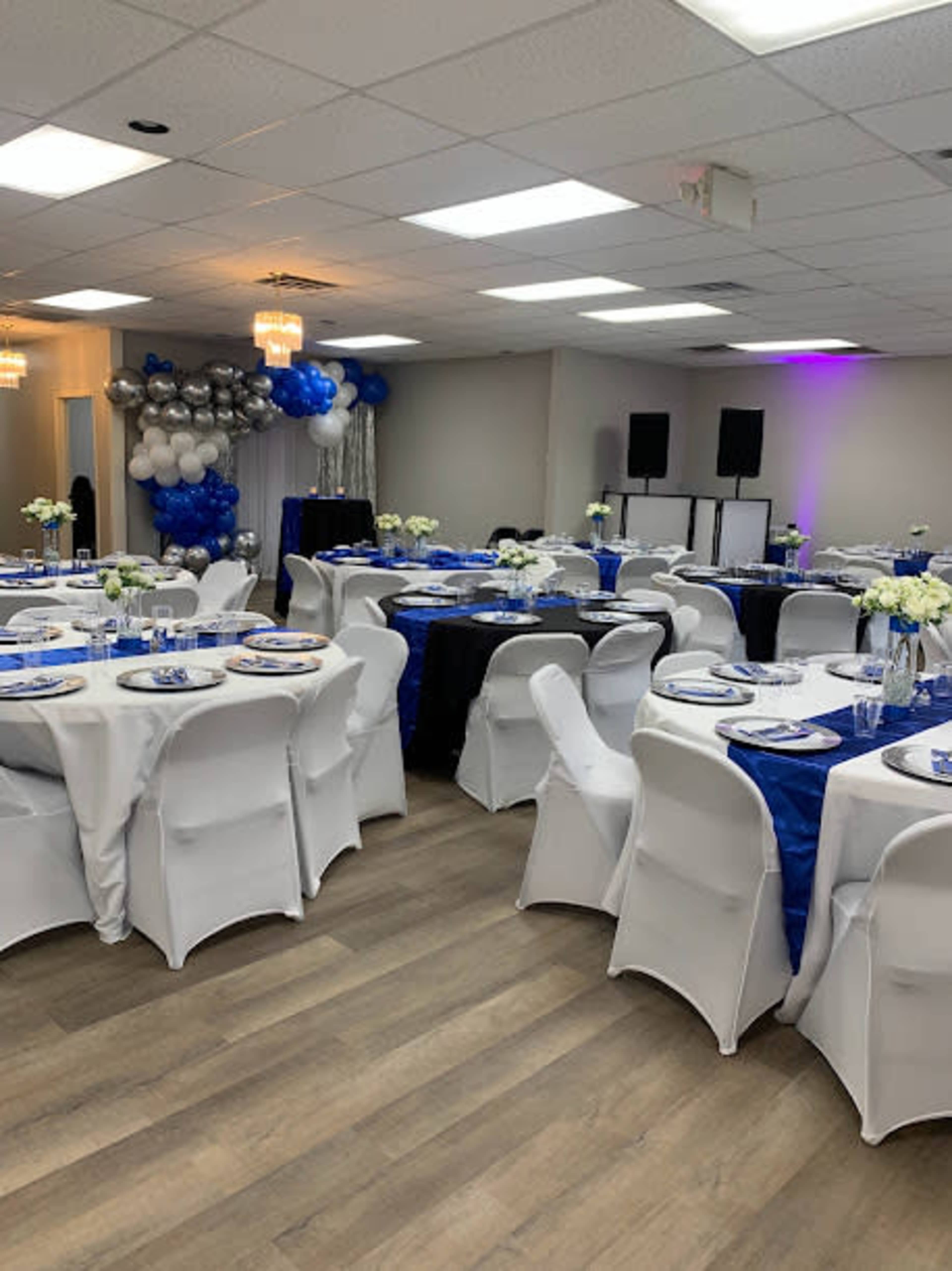 A decorated event space with tables set for dining, featuring white and blue tablecloths, floral centerpieces, and balloon decorations.