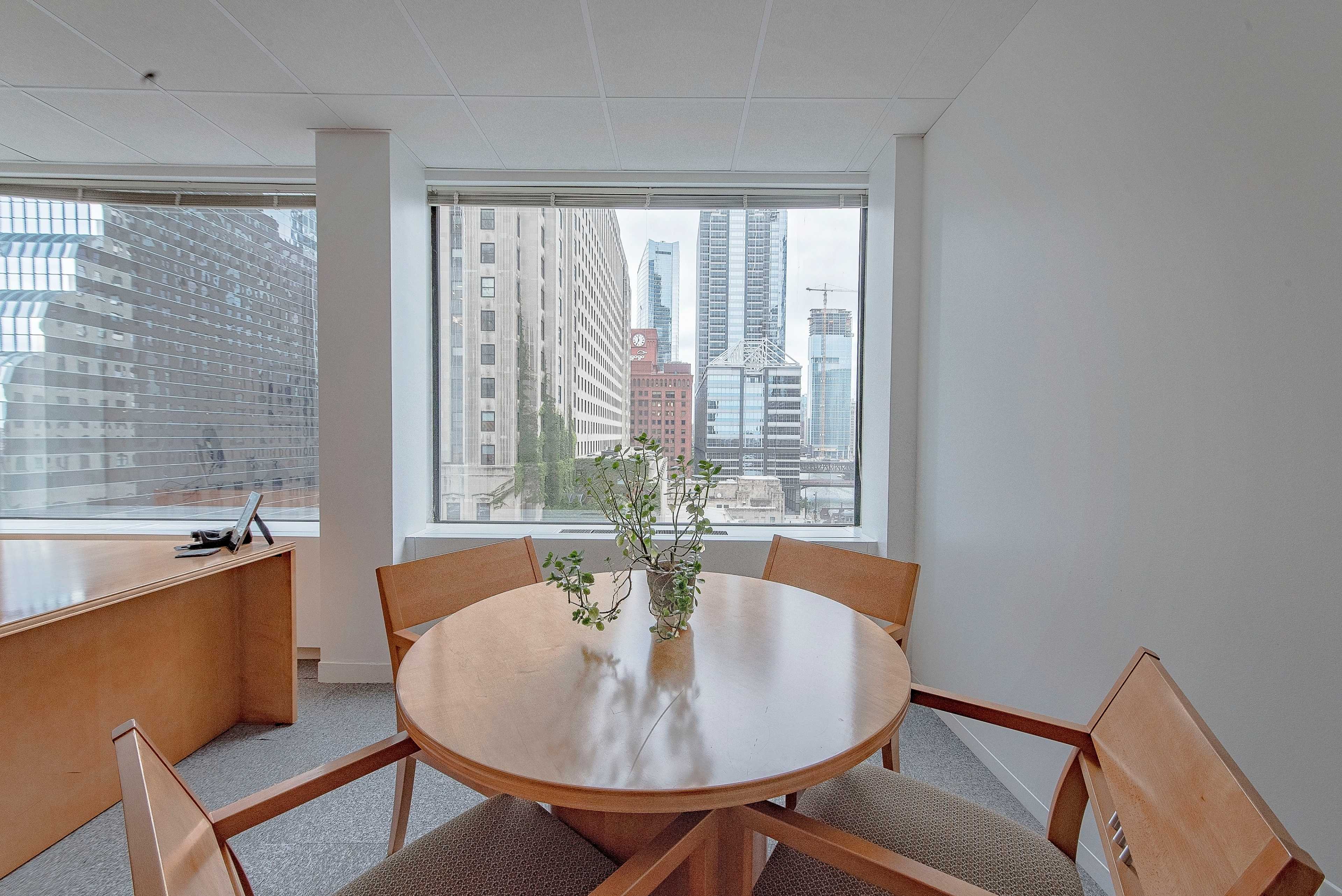 A small round table with wooden chairs is set near a window overlooking a cityscape featuring tall buildings and construction sites.