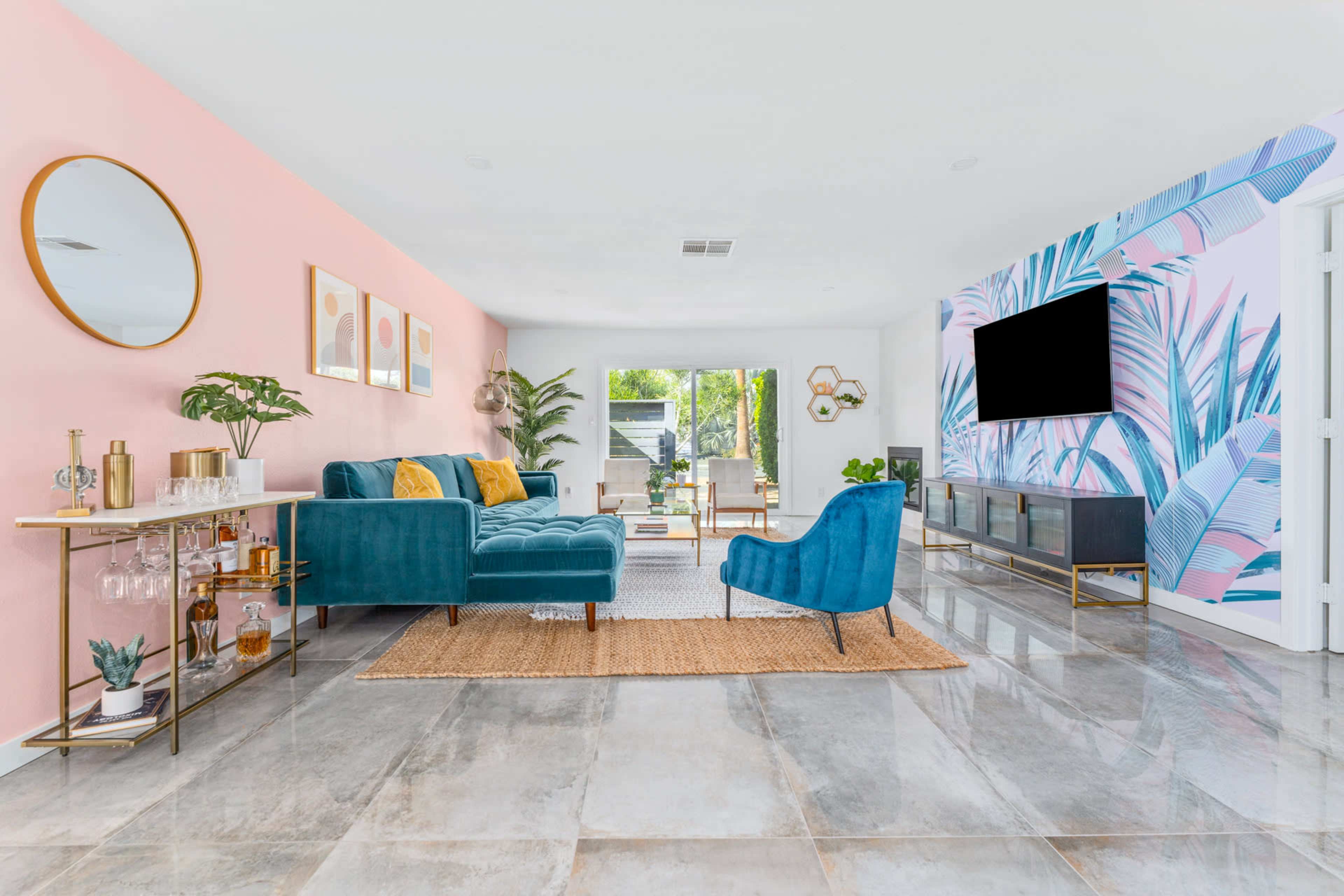 A modern living room features a blue velvet sectional sofa, a round mirror, tropical wall art, and a large TV against a pastel wall, with natural light streaming through large windows.