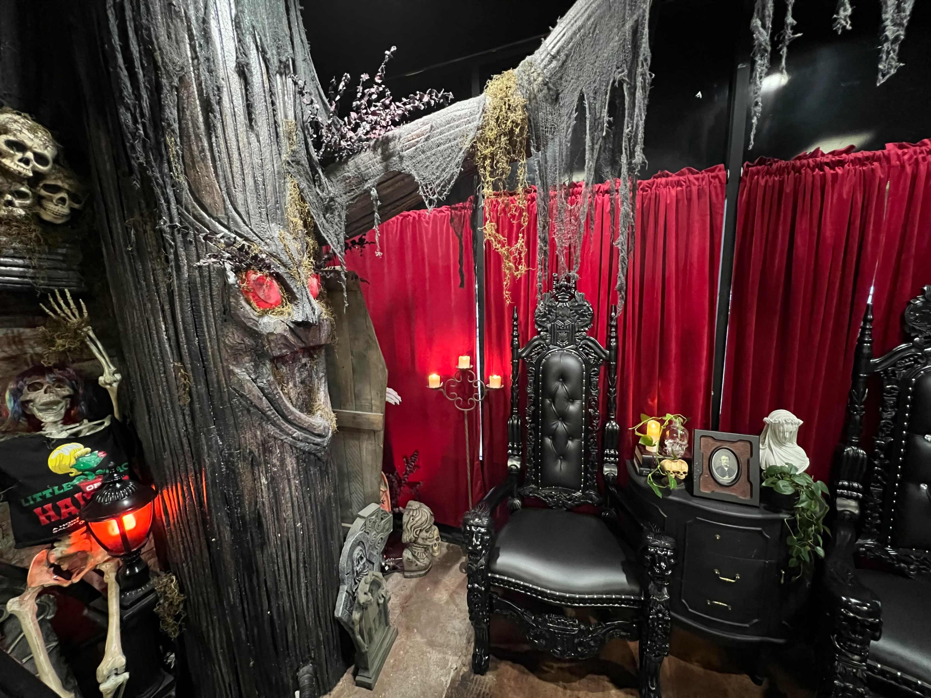 The scene features a dark, elaborate room with a large, gnarled tree, two ornate black chairs, red drapes, and eerie decorations including skulls and skeletons.