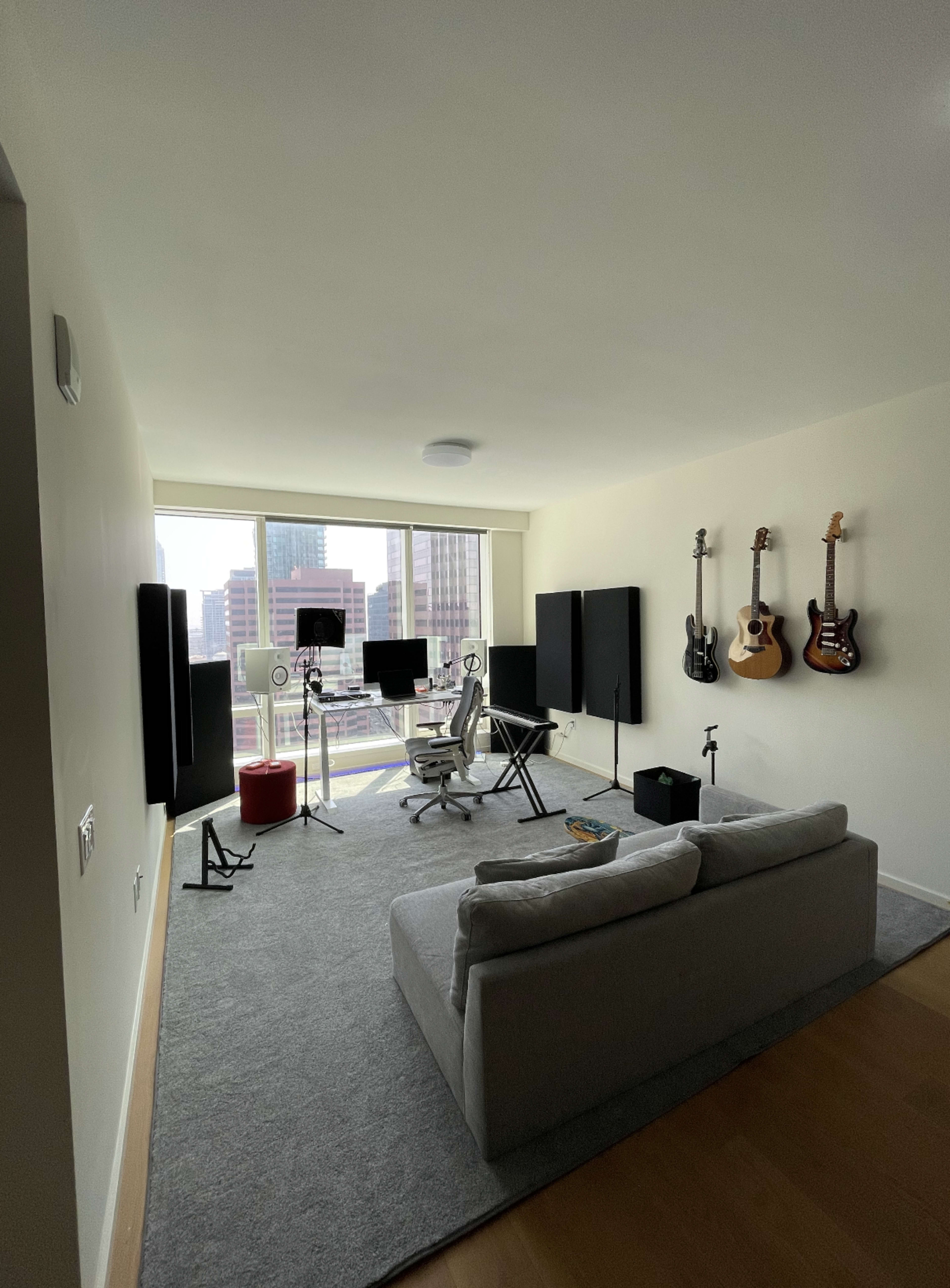 A modern living space features a gray sofa, an office setup with multiple monitors, and three guitars mounted on the wall.