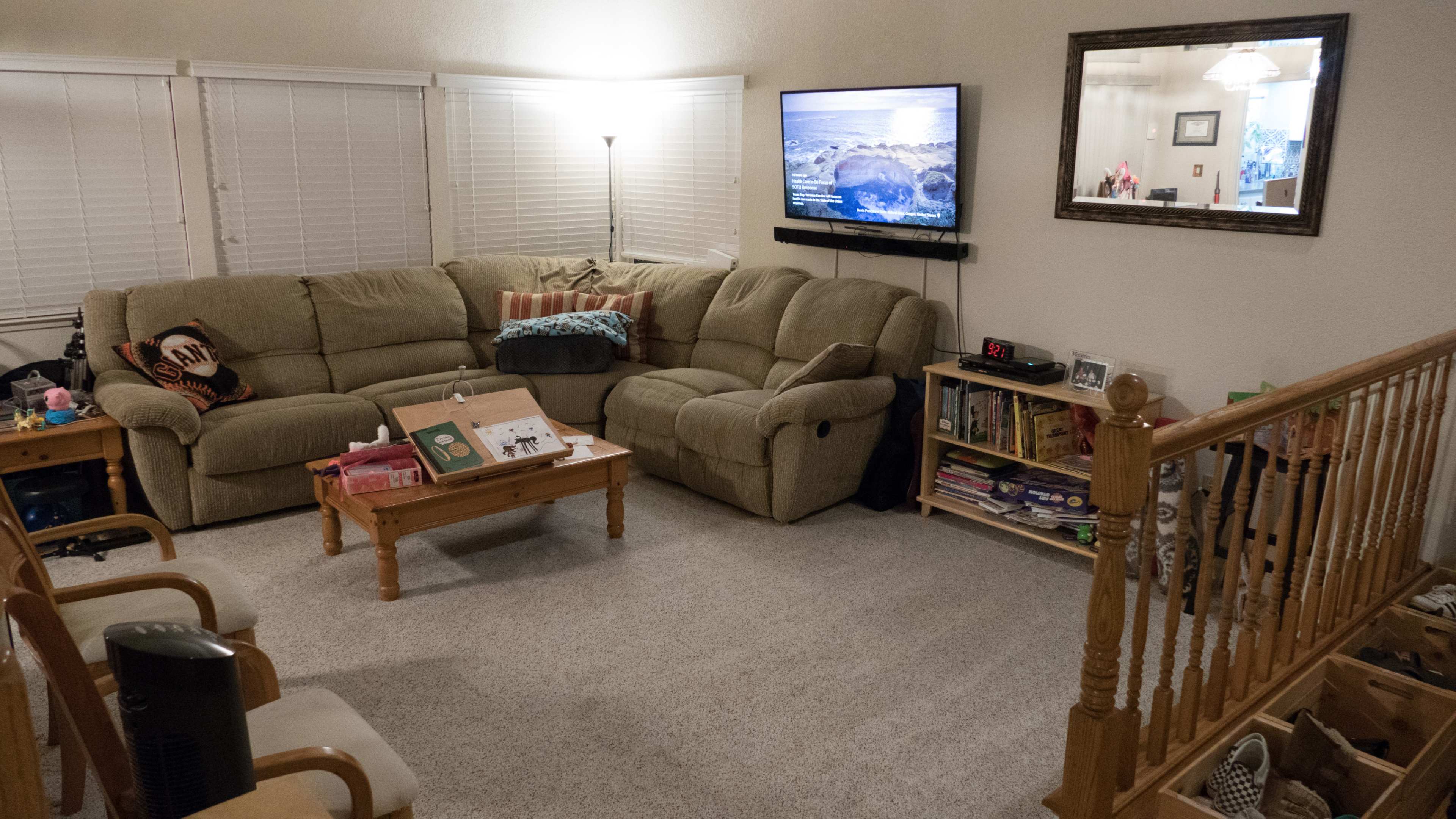 The living room features a sectional couch facing a wall-mounted television, with a wooden coffee table and a bookshelf full of books in the corner.