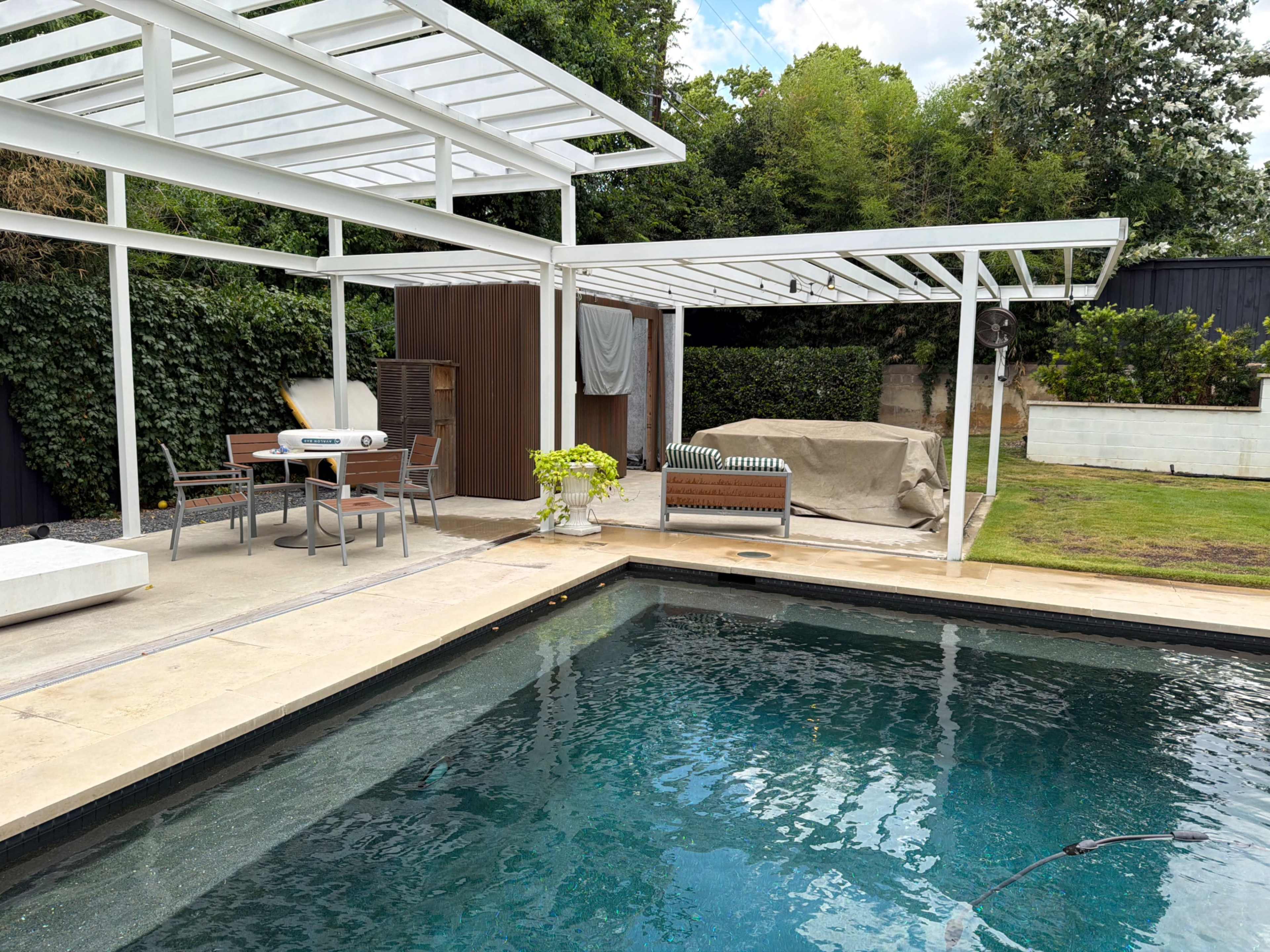 The image shows a backyard with a swimming pool, a covered seating area, and a partially covered hot tub.