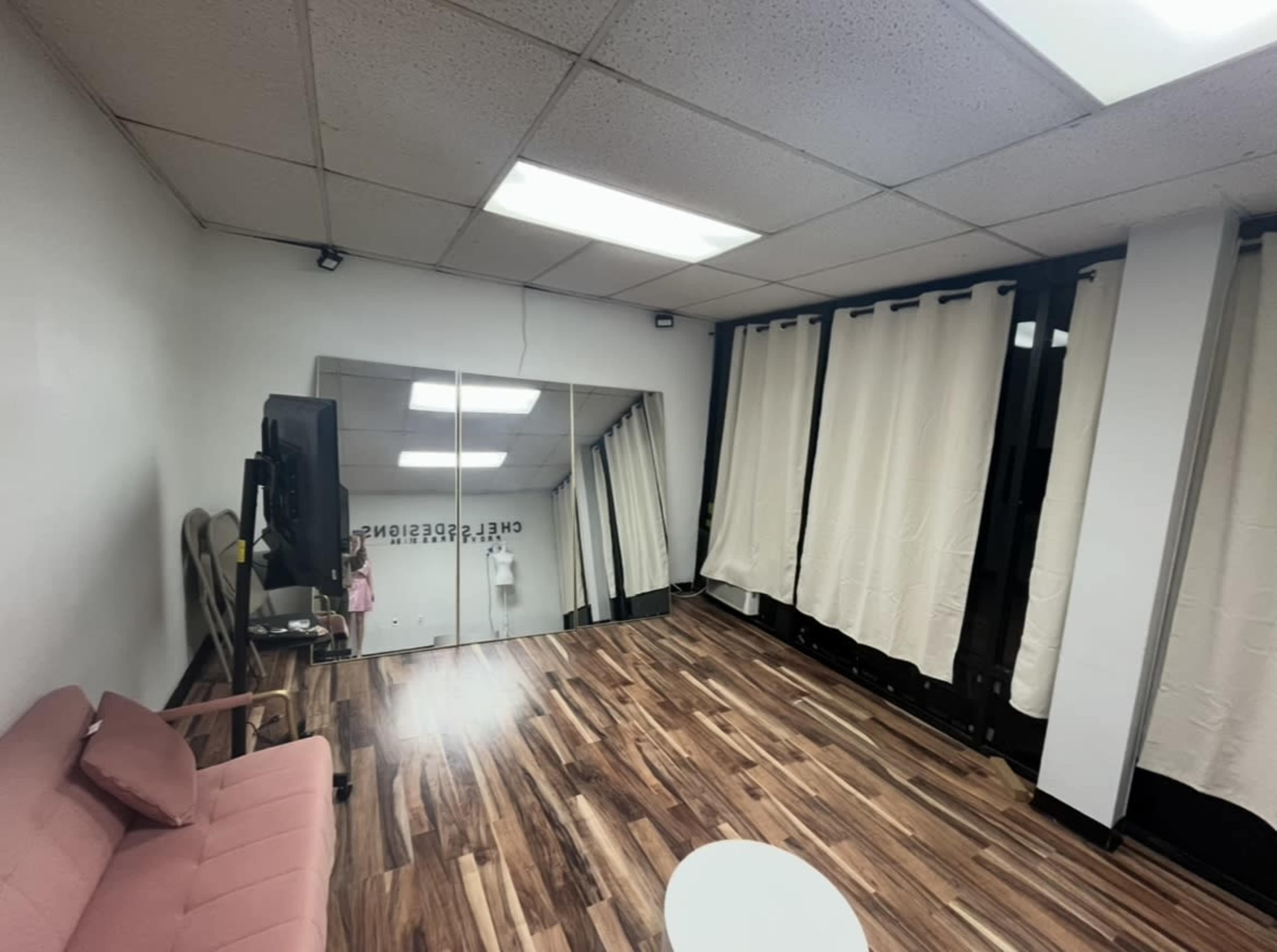 The image shows a small, sparsely furnished room with a pink couch, a television, a mirror on the wall, and sheer curtains covering the windows.