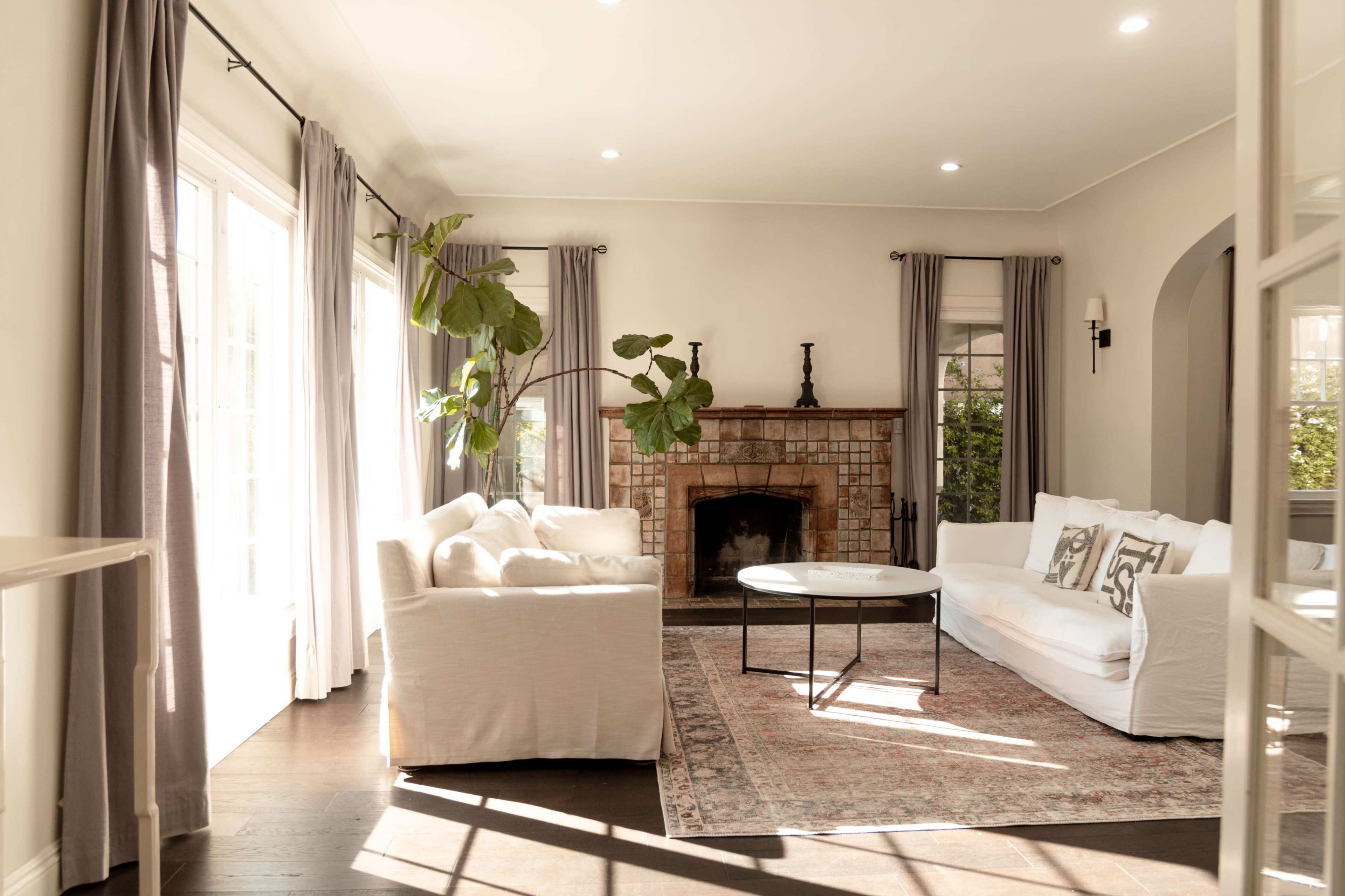 A bright living room features a white sofa, a round coffee table, and a brick fireplace, complemented by large windows and curtains.