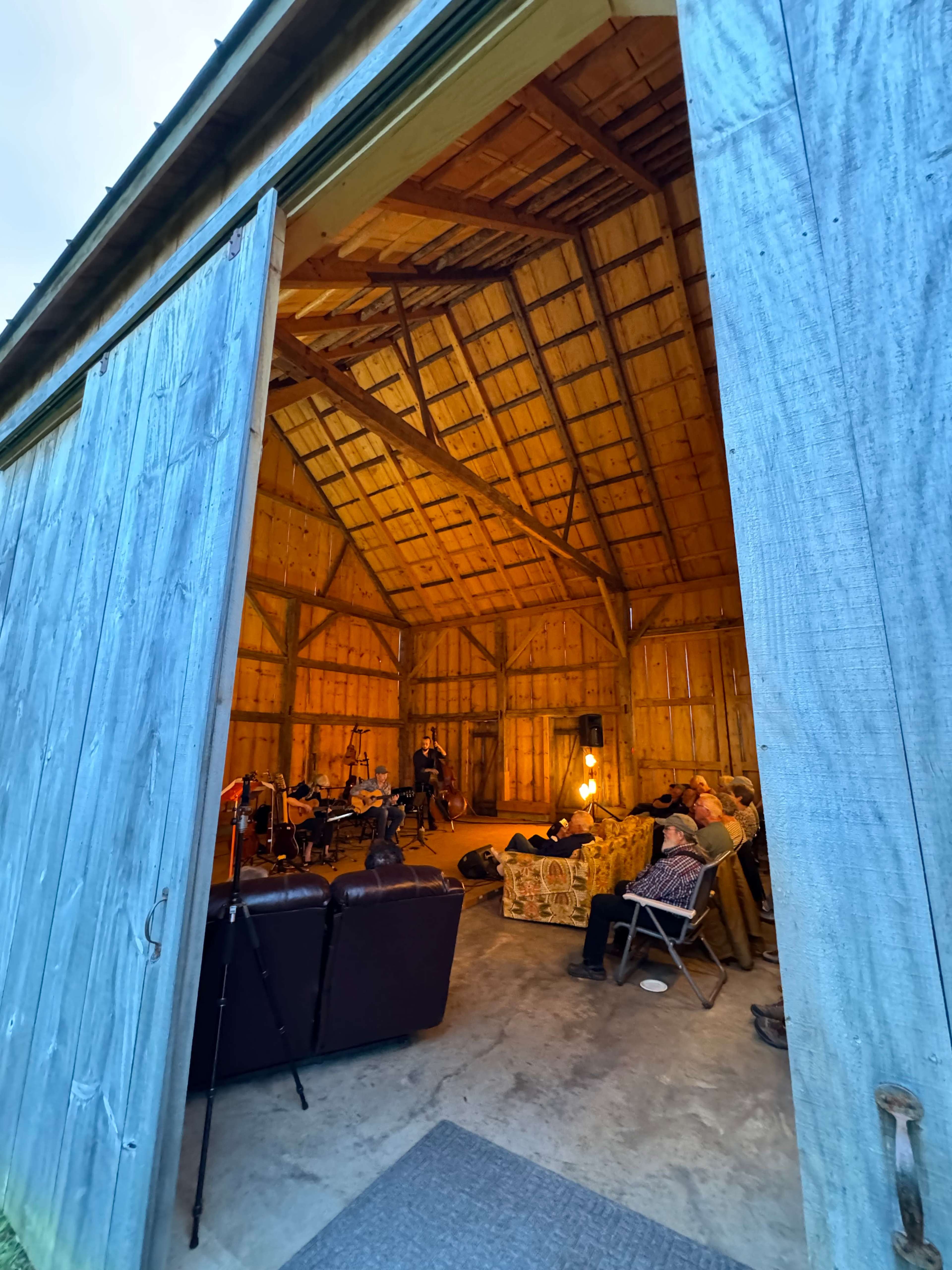A partially open barn door reveals a gathering inside, with people seated on couches and chairs under warm lighting.