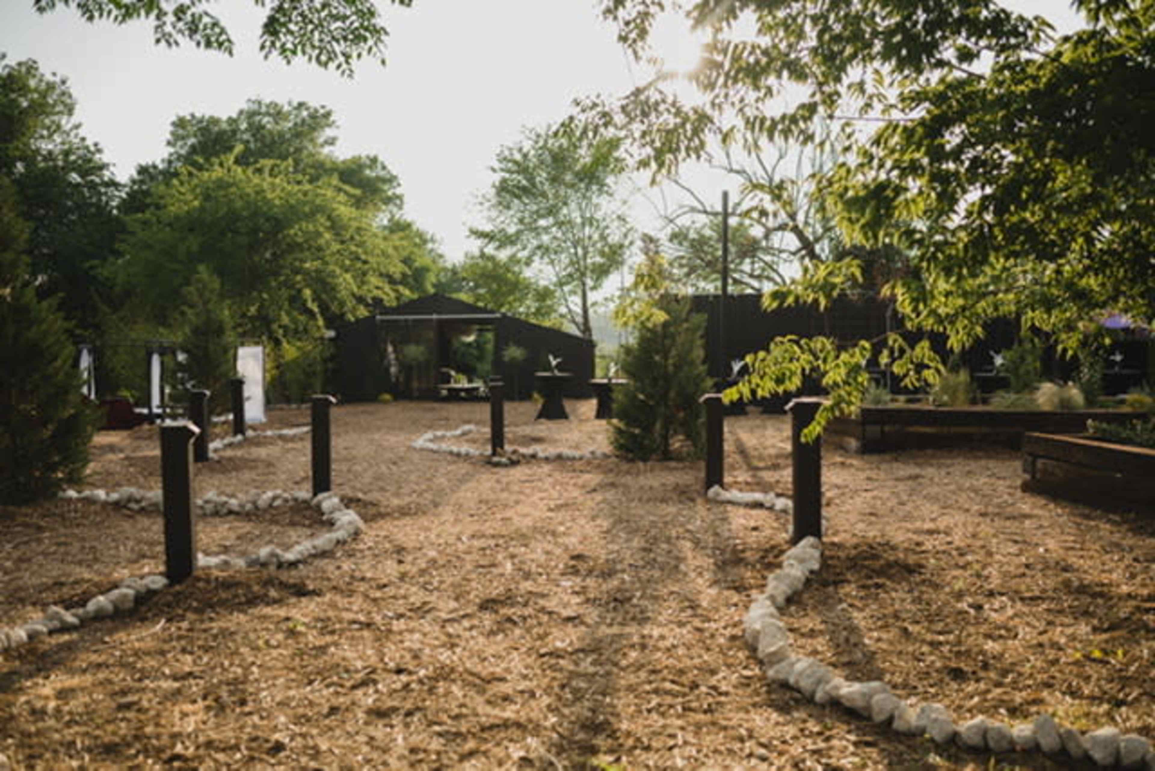 The image shows a landscaped outdoor area with gravel pathways bordered by stones and surrounded by trees and garden features.