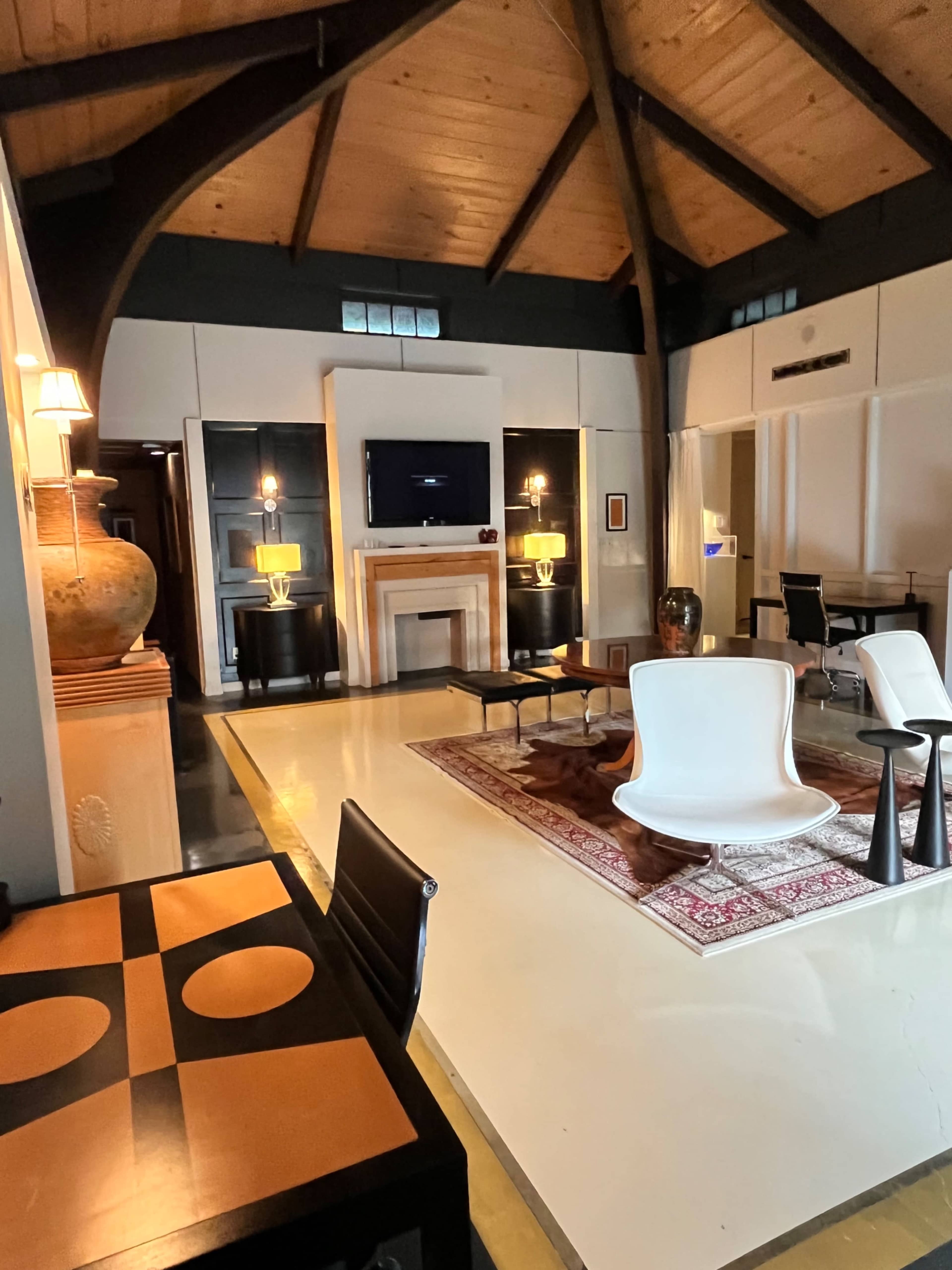 The image shows a modern living space with high wooden ceilings, a decorative fireplace, and contemporary furniture arranged around a patterned rug.
