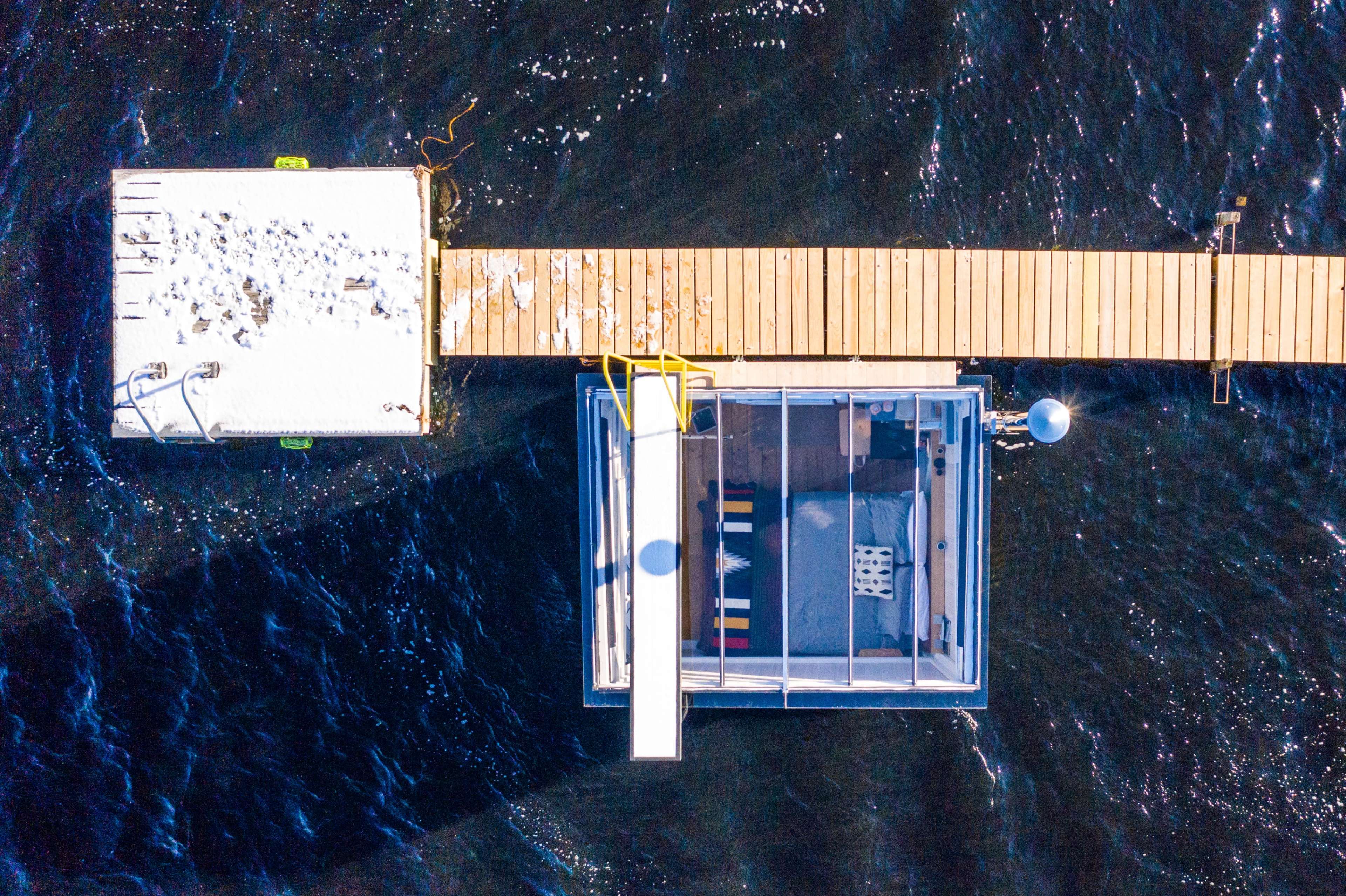 An aerial view shows a wooden dock leading to a glass-enclosed structure on dark water.