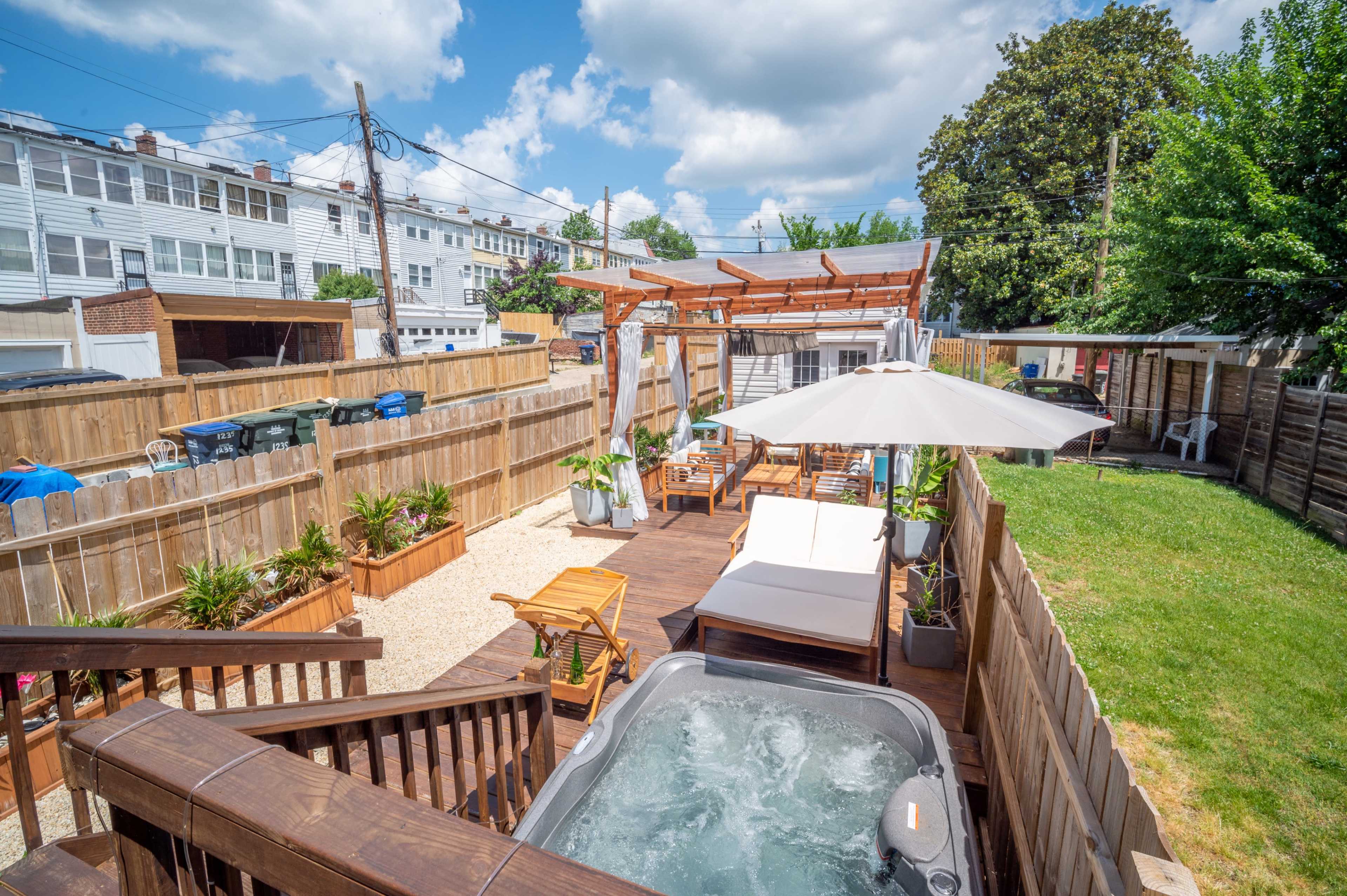 A backyard with a hot tub, a shaded seating area, potted plants, and a wooden deck surrounded by fences and houses in the background.