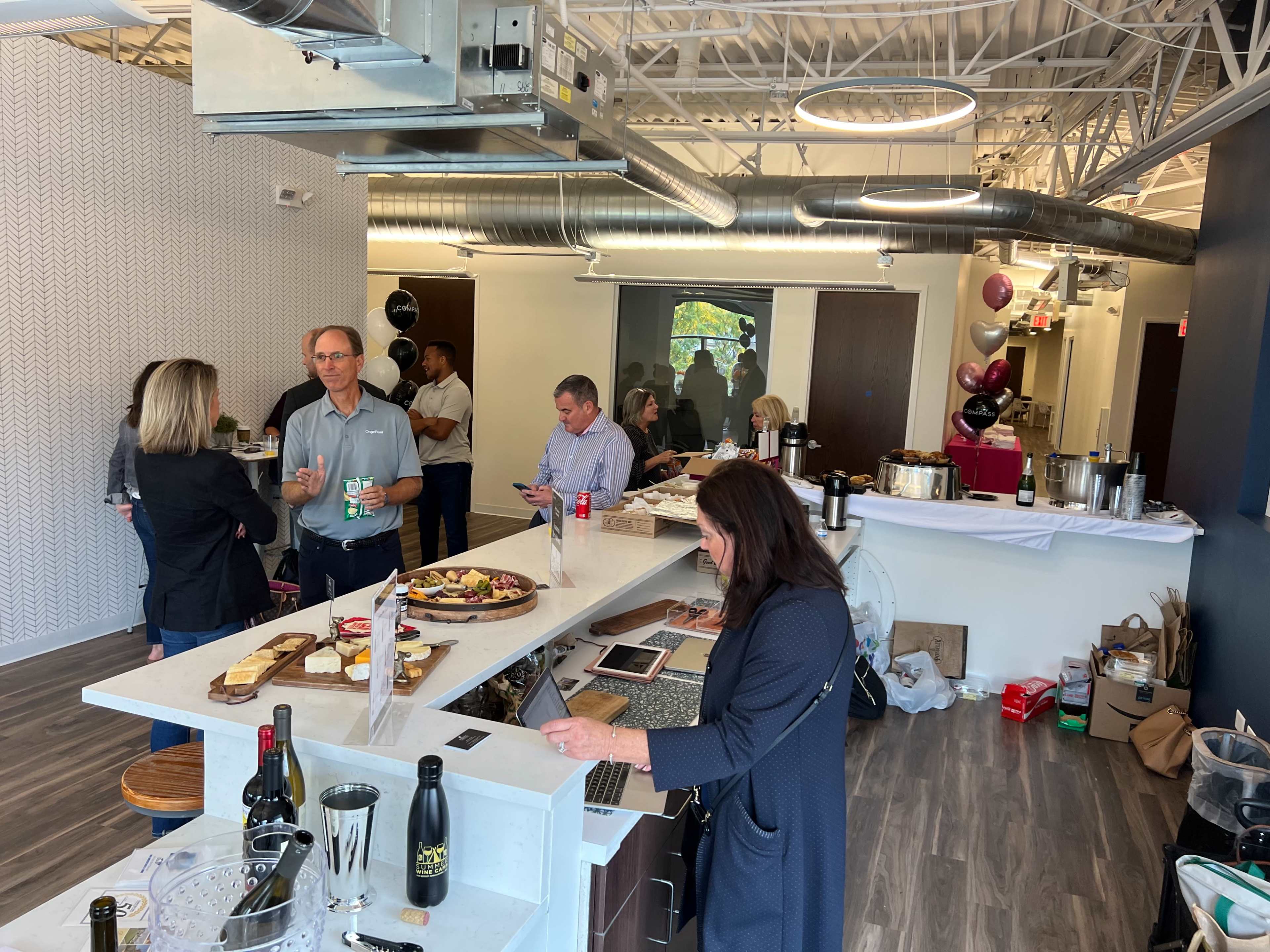 A group of people socialize in an office kitchen area, with food and drinks laid out on a counter.