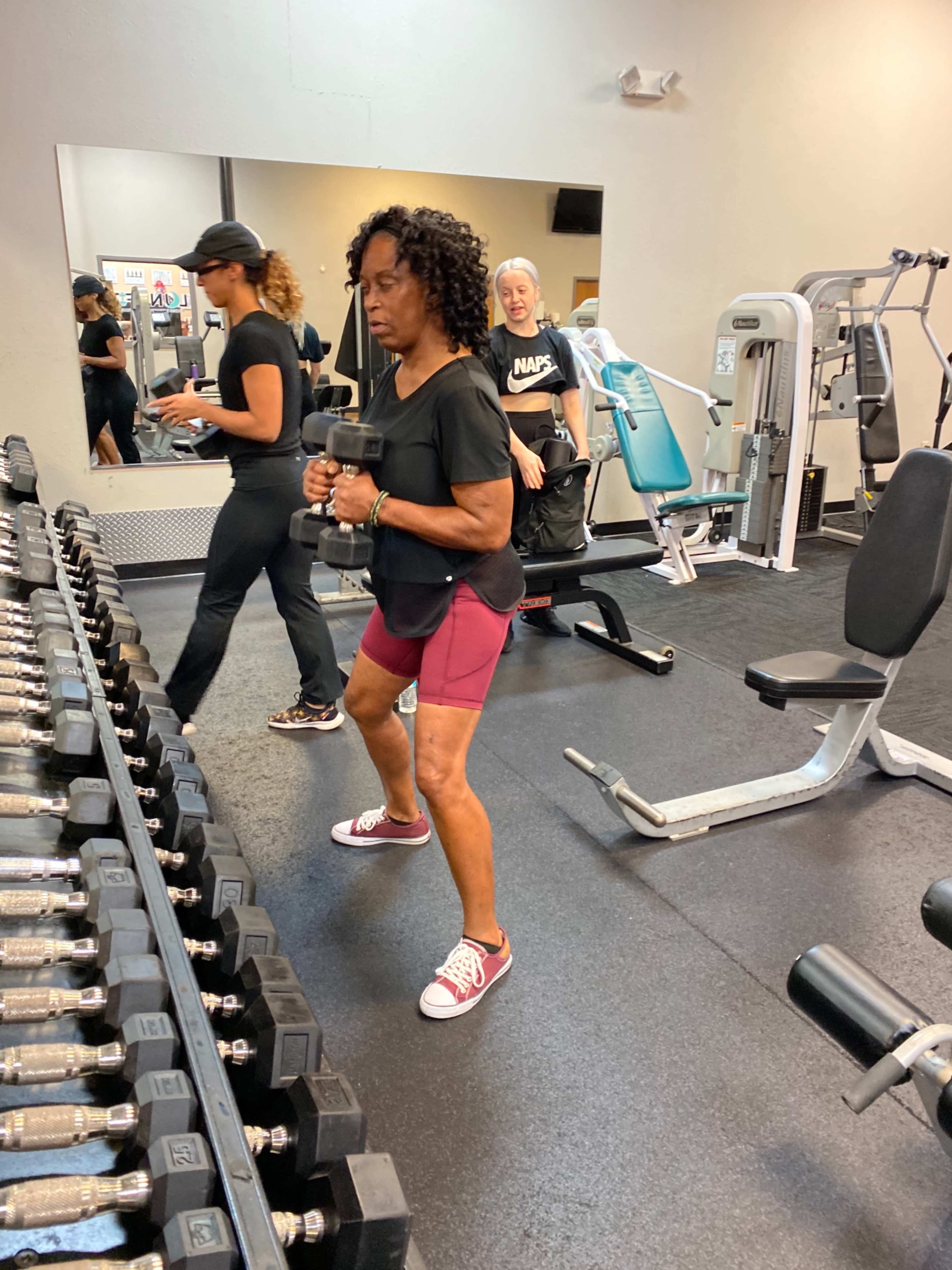 A woman in a black shirt and maroon shorts is performing a weighted exercise with dumbbells in a gym, while another woman and a third figure are engaged in different activities nearby.