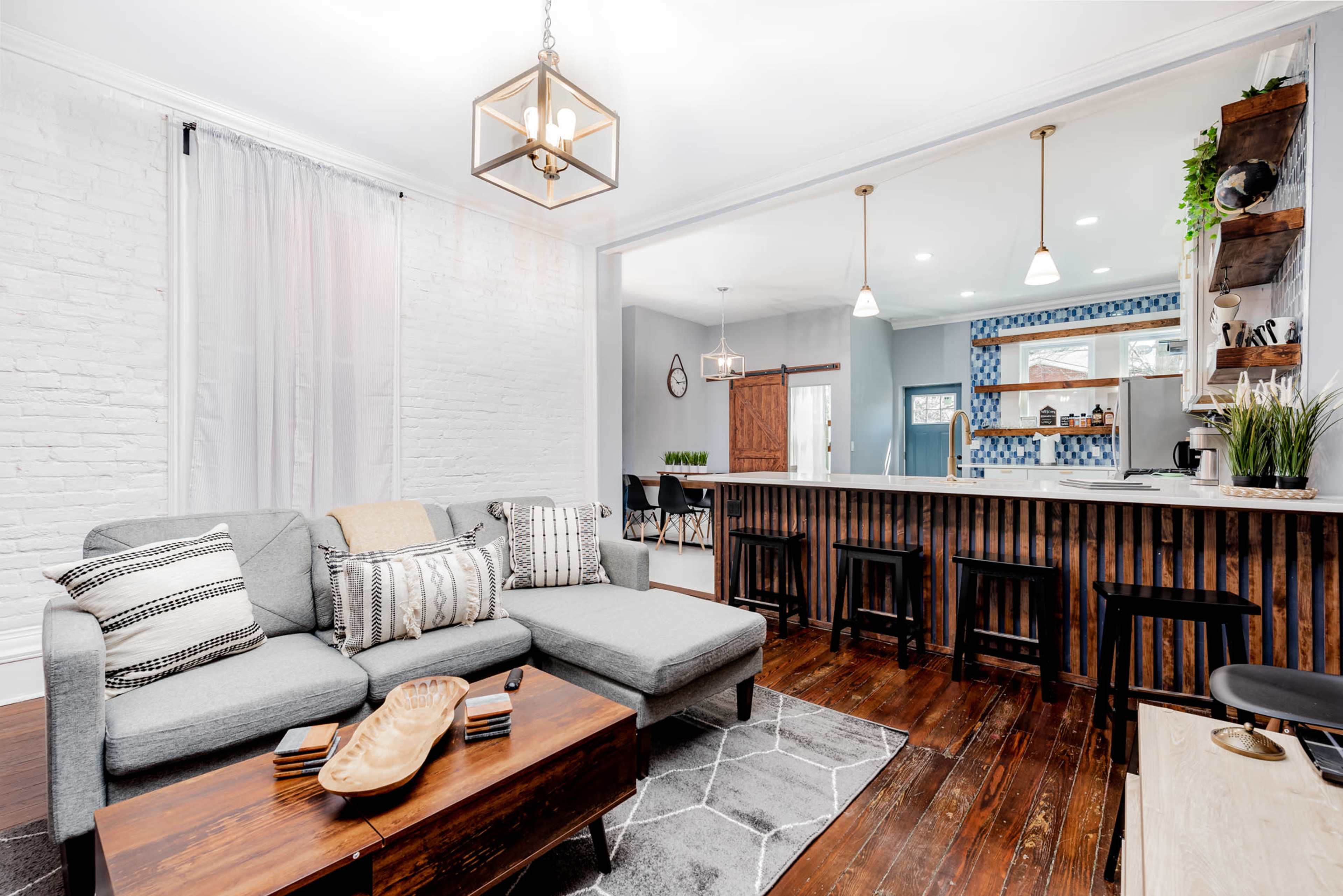 A modern living room features a gray sectional sofa, a wooden coffee table, and an open kitchen with a bar and blue tile backsplash.