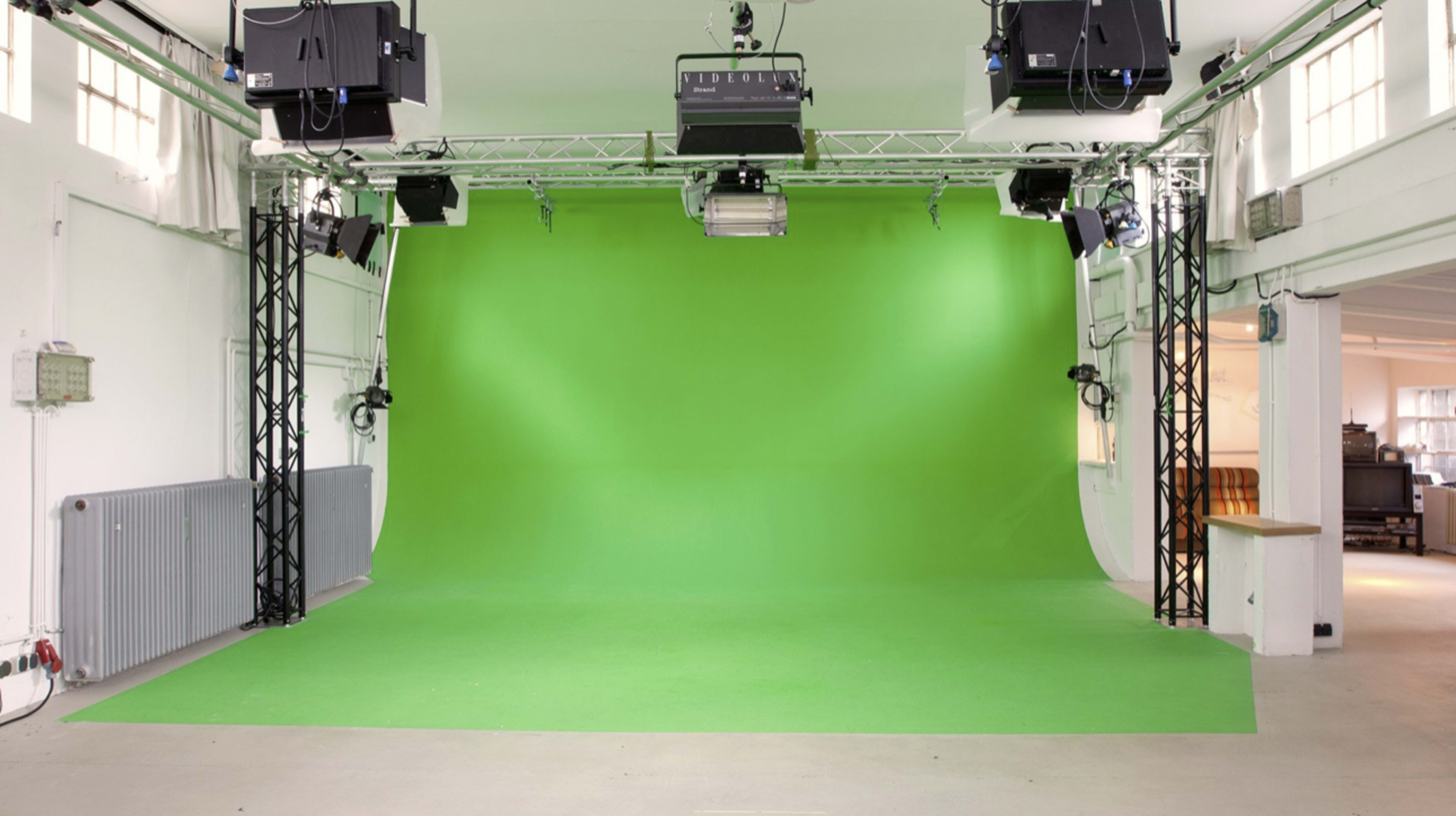 The image shows a green screen studio with a seamless green backdrop and overhead lighting equipment.