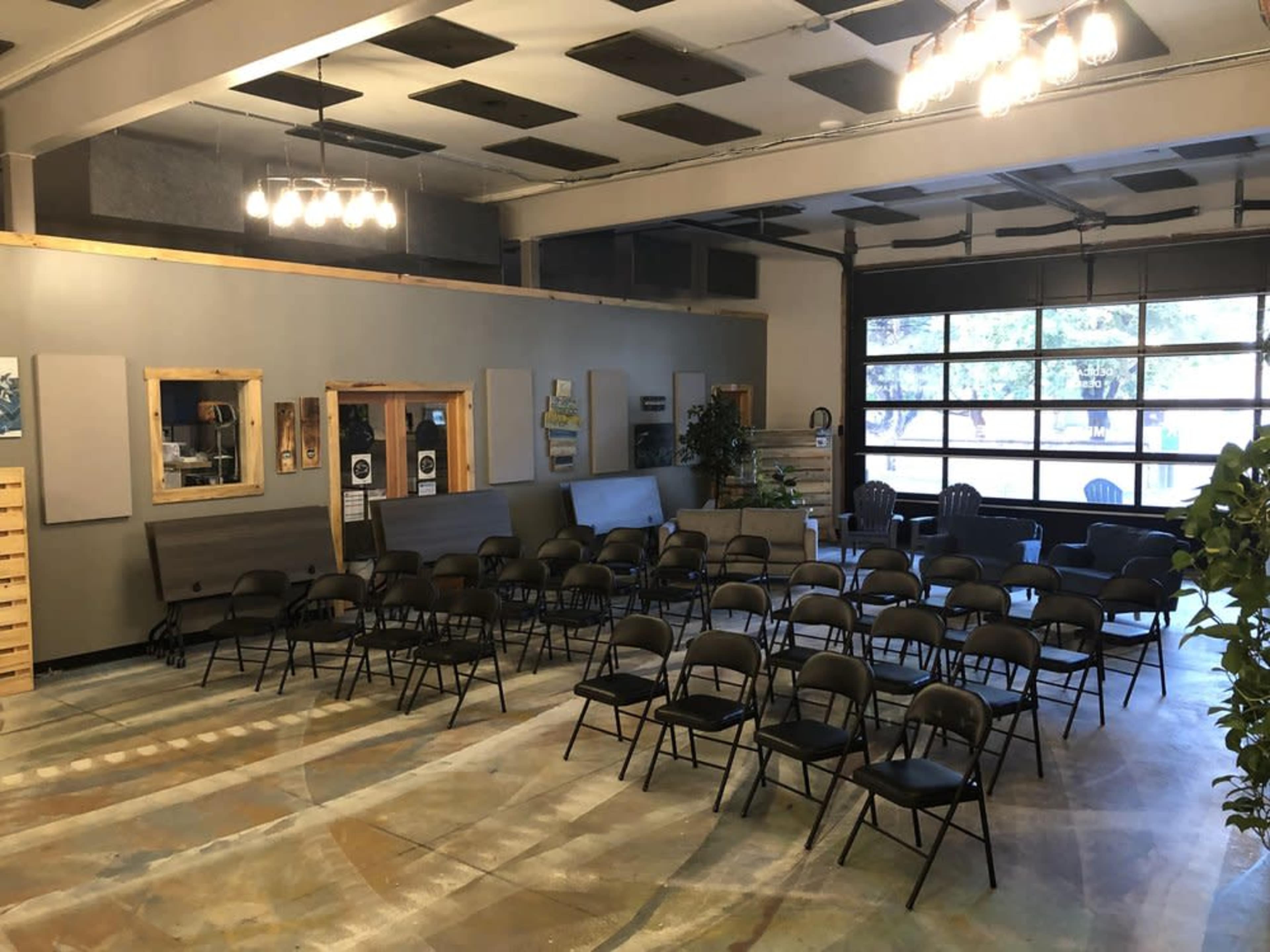 A room set up with rows of black folding chairs facing a blank wall, with large windows allowing light to enter.