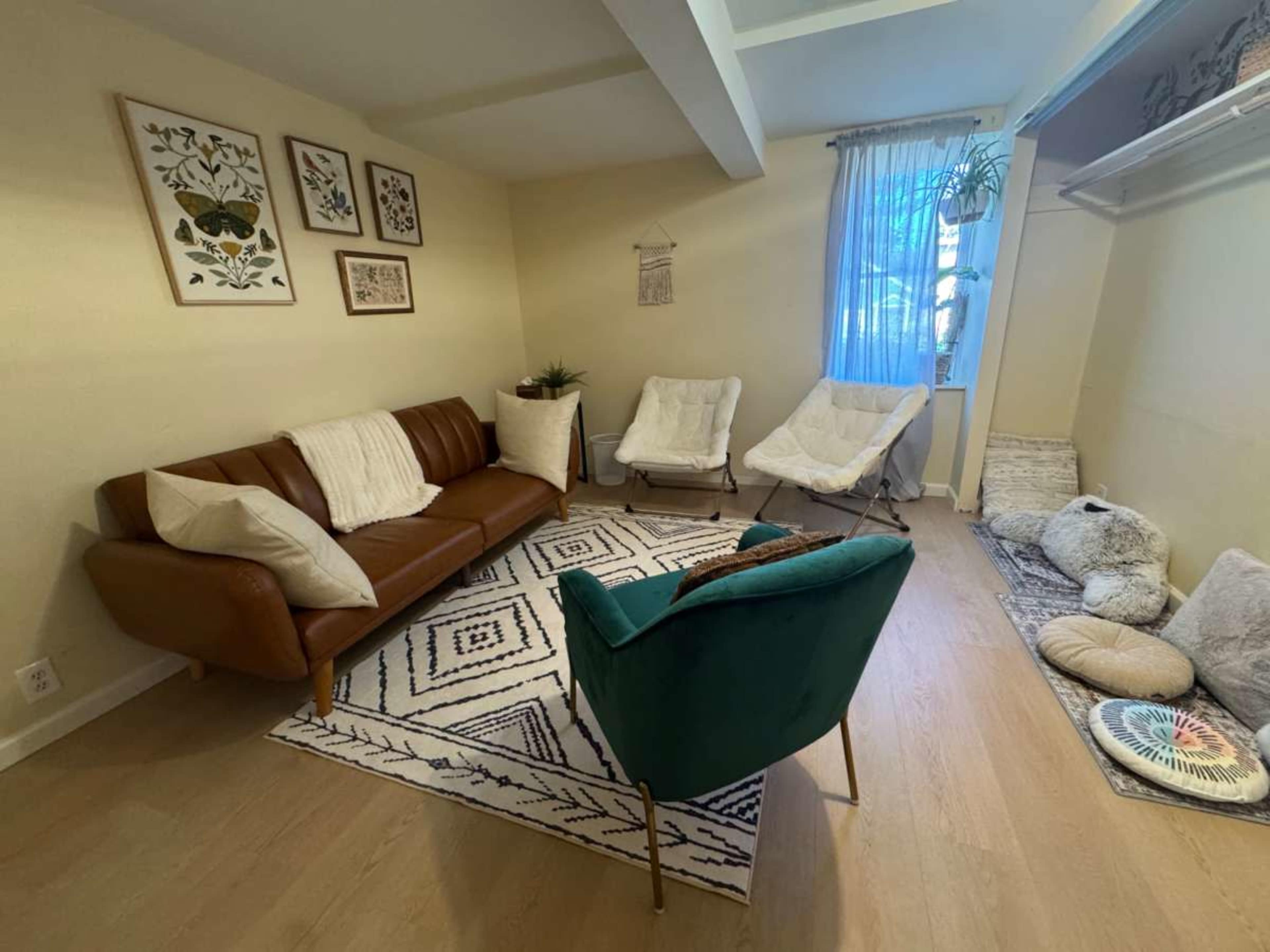 The image shows a cozy living room with a brown sofa, two white armchairs, and a patterned area rug, complemented by wall art and natural light from a window.