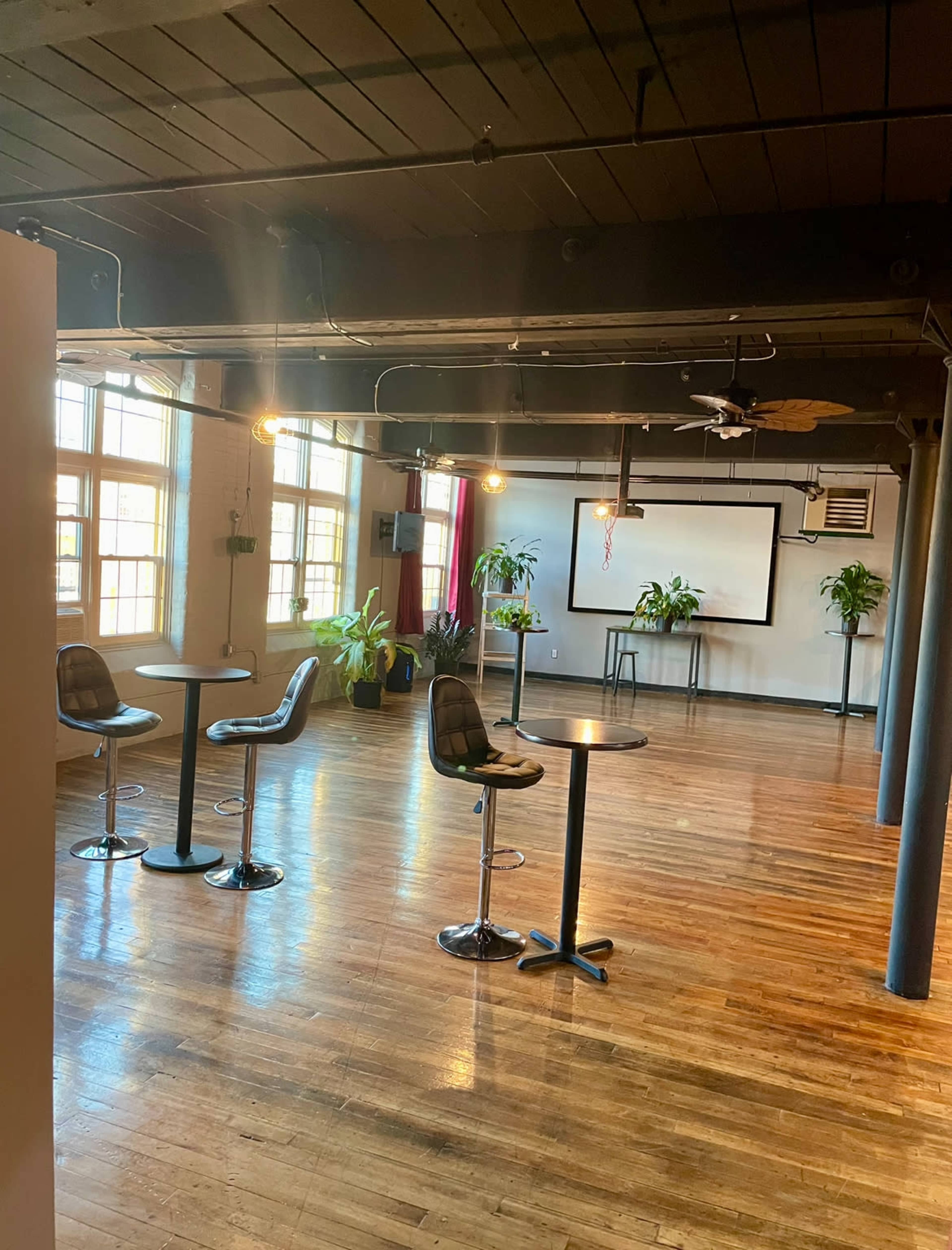 The image shows a spacious room with a polished wooden floor, featuring bar stools and tables, plants, and a large screen at the front.